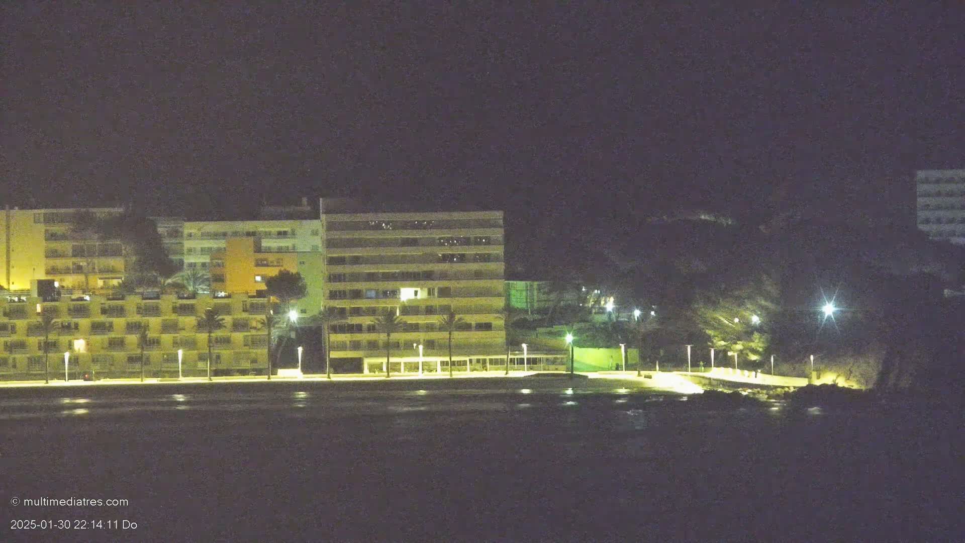 A row of brightly lit buildings stands along a dark shoreline at night.