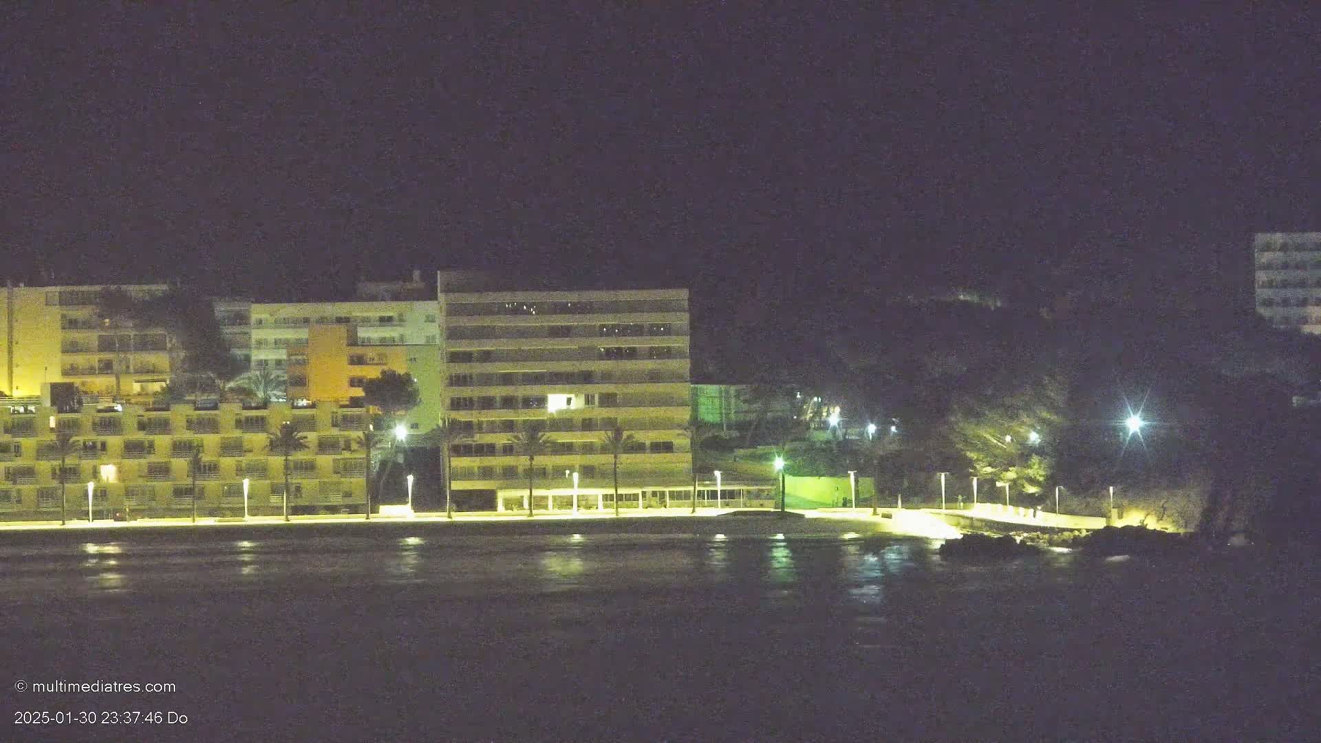 A row of illuminated buildings stands along a dark shoreline at night, with calm water in the foreground.