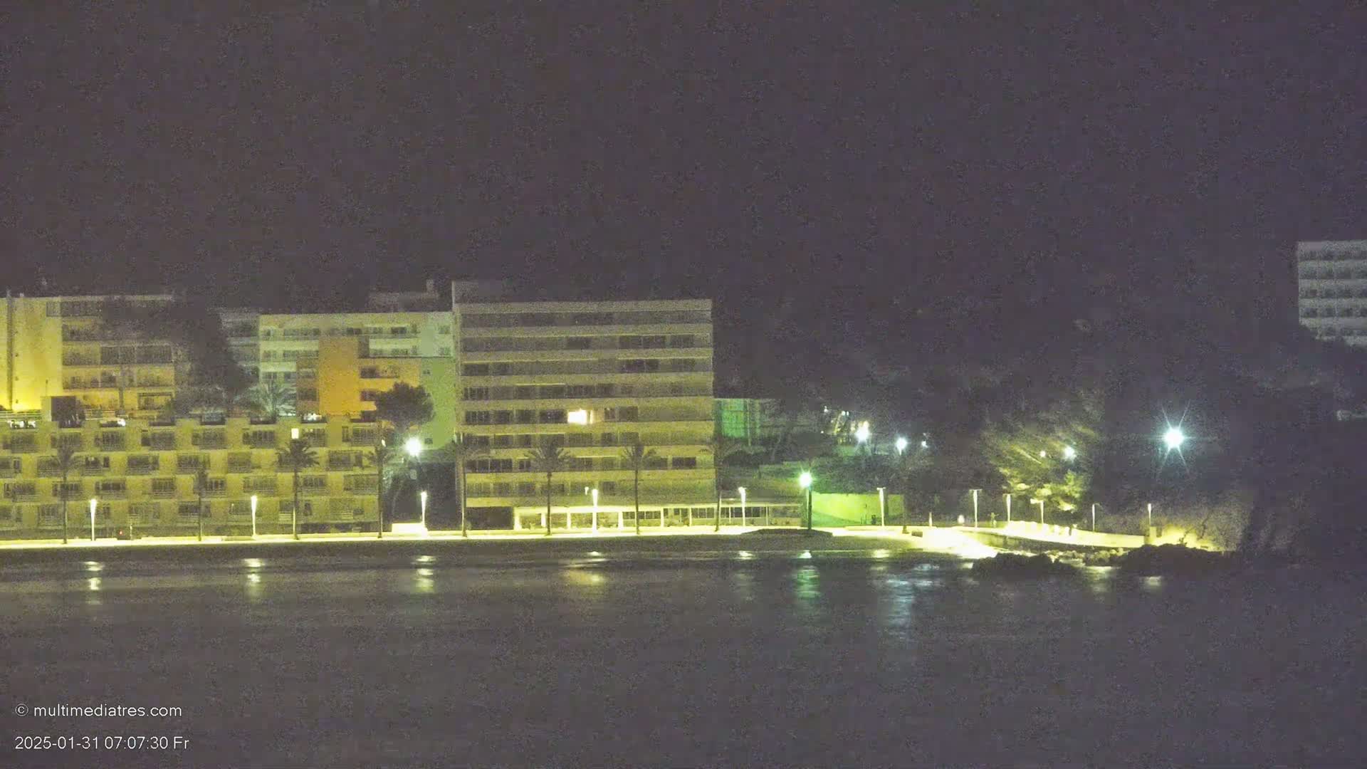 A line of illuminated beachfront buildings stands beside a dark body of water under a clear night sky.