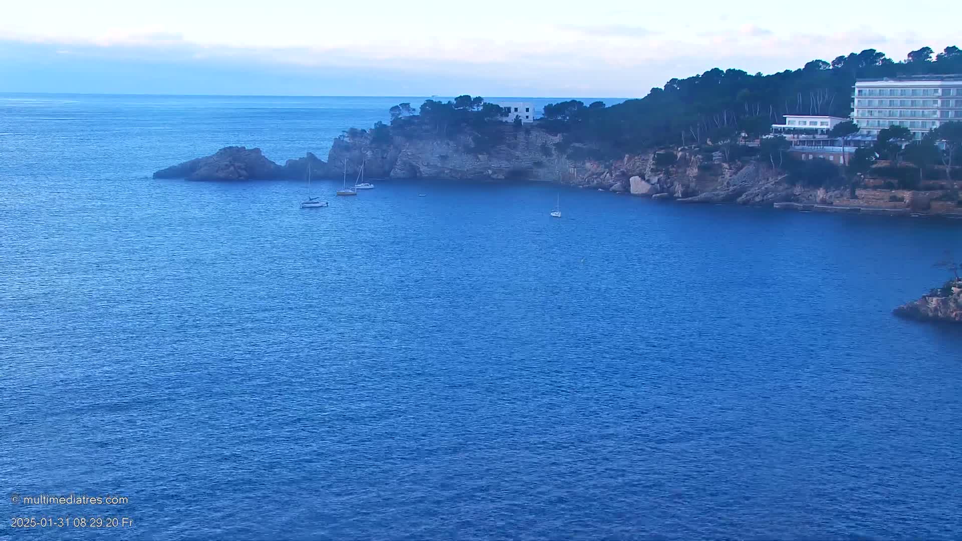 A calm, blue sea laps against a rocky coastline dotted with a few sailboats and a large building nestled among the trees.