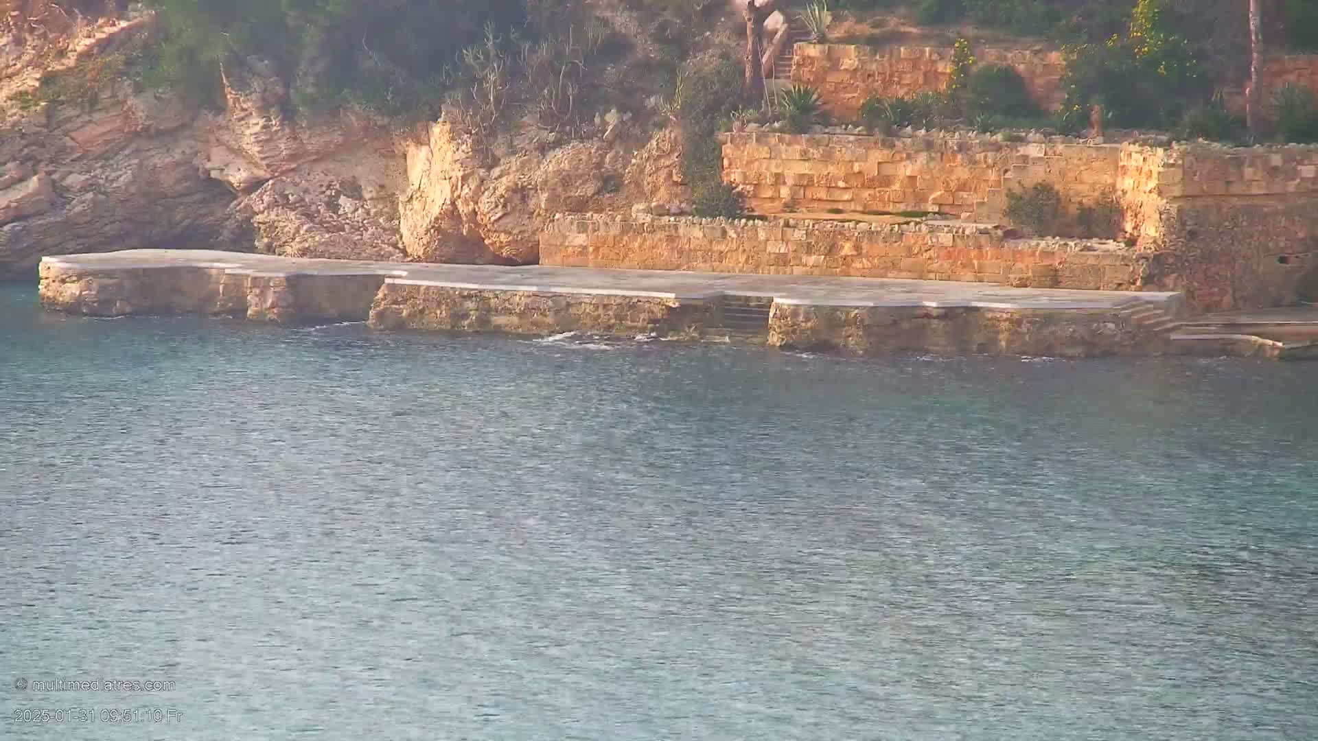 A stone platform extends from a rocky, terraced hillside into calm, blue-green water.