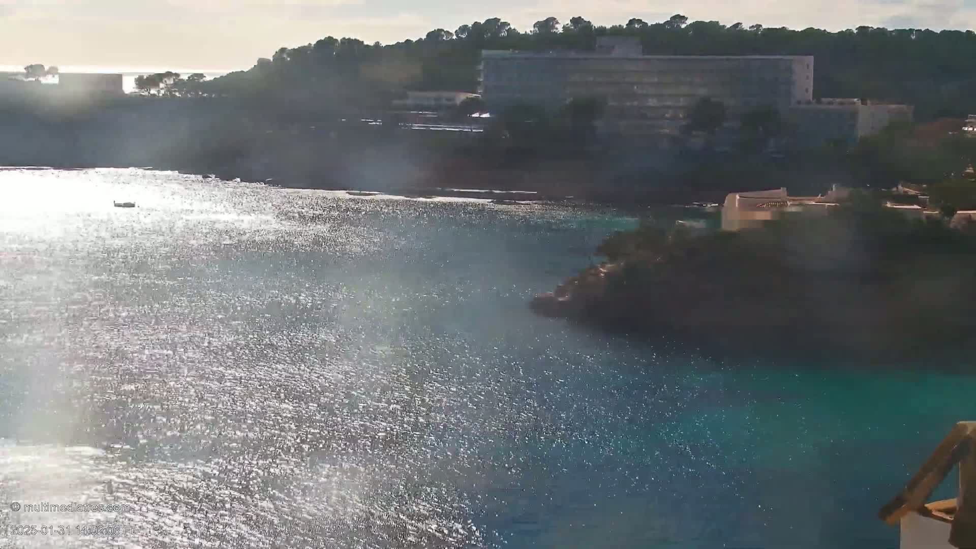 Sparkling water reflects sunlight on a calm, sunny day near a large hotel nestled among green hills.
