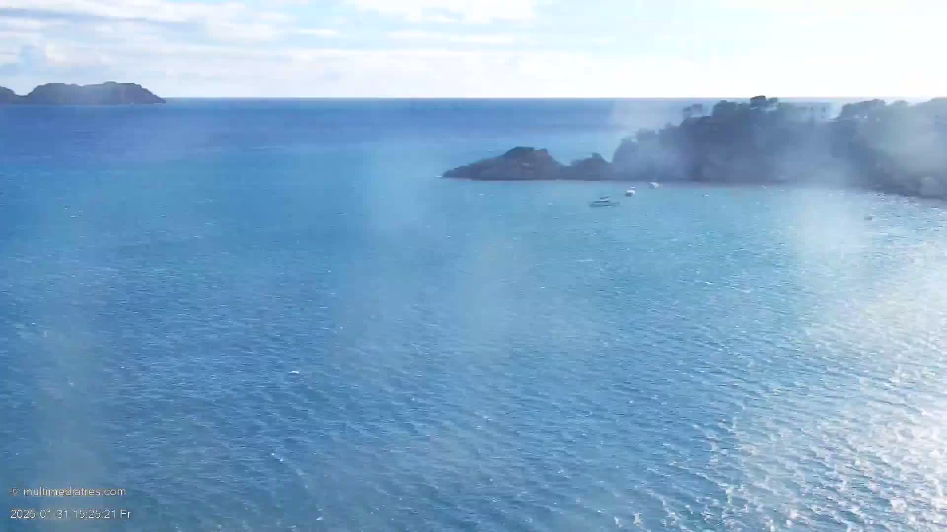 A hazy, sunny day shows a calm blue sea with small boats near rocky islands and a larger landmass in the distance.