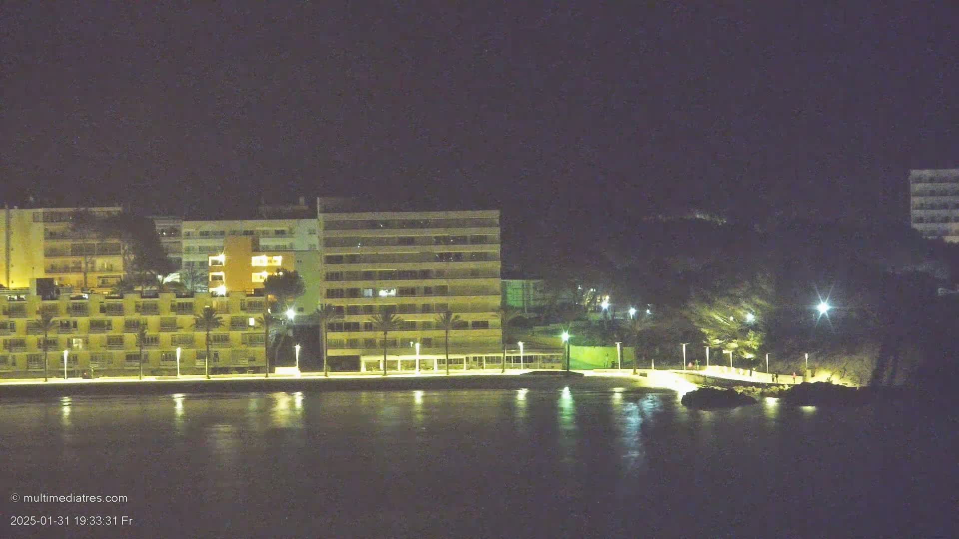 A row of illuminated buildings lines a waterfront at night, reflected in the calm water.