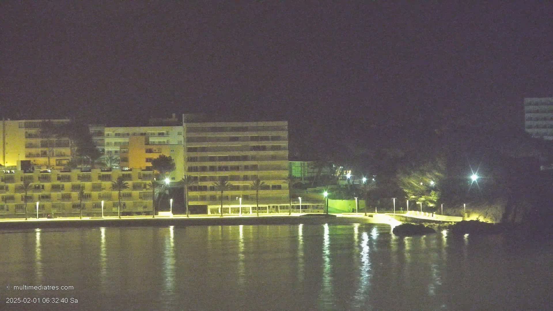 At night, several multi-story buildings are illuminated along a waterfront, with their lights reflecting on calm water.