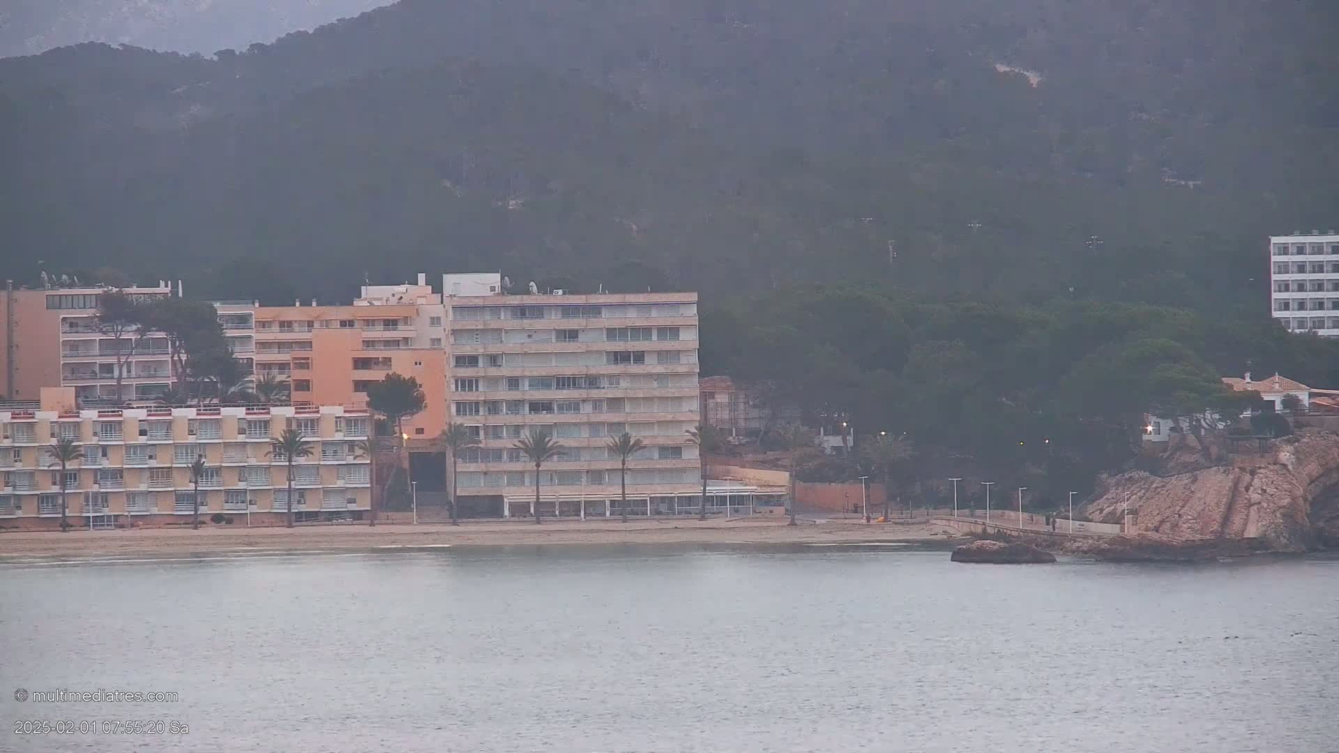 A hazy view of several multi-story buildings situated on a beach next to a calm body of water and a rocky outcrop, with a mountain range in the background.