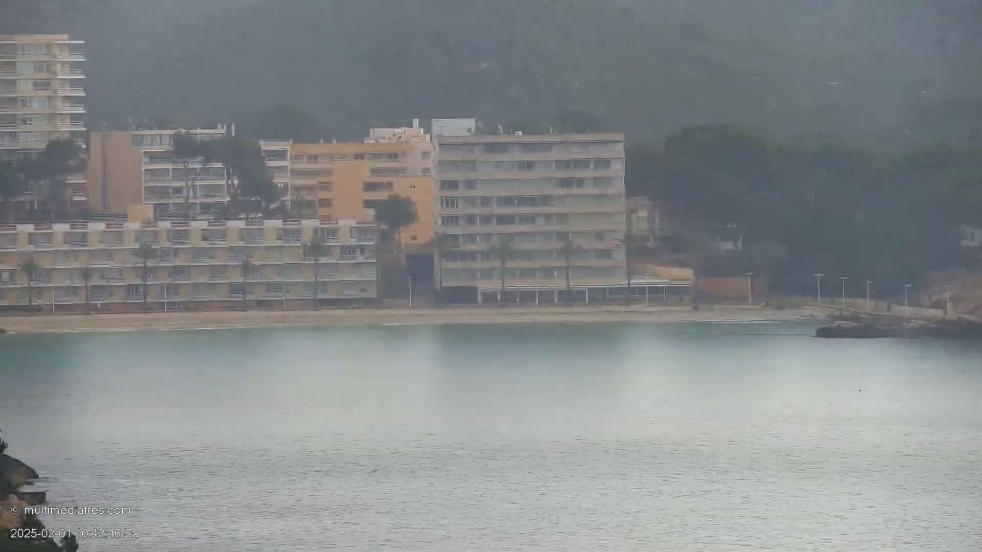 A hazy view shows several multi-story buildings on a beach next to calm, gray water.