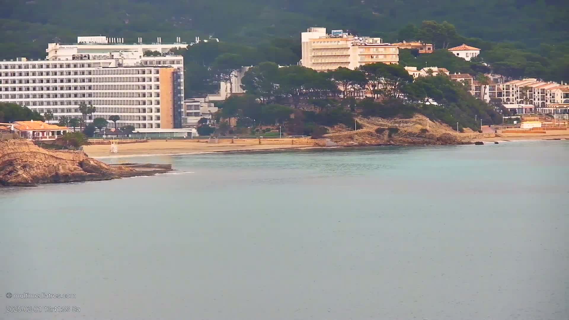 A sandy beach with hotels and buildings on a hillside overlooks calm, light-blue ocean water under an overcast sky.