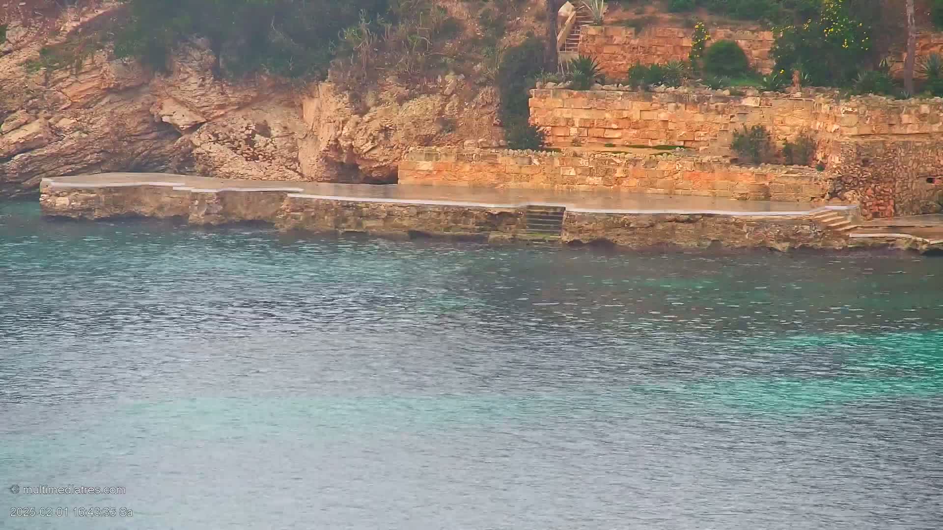 A stone platform with stairs leading into calm, clear teal water sits against a rocky cliffside.