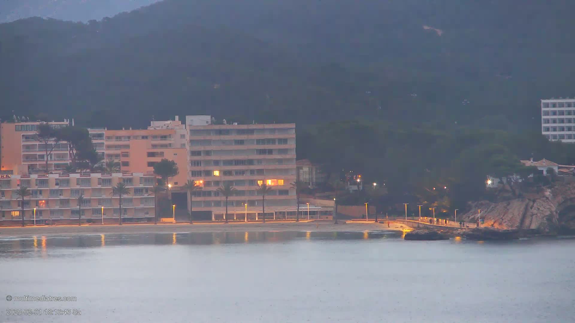 A row of low-rise buildings with lights on illuminates a beach at dusk under a hazy sky, with calm water in the foreground.