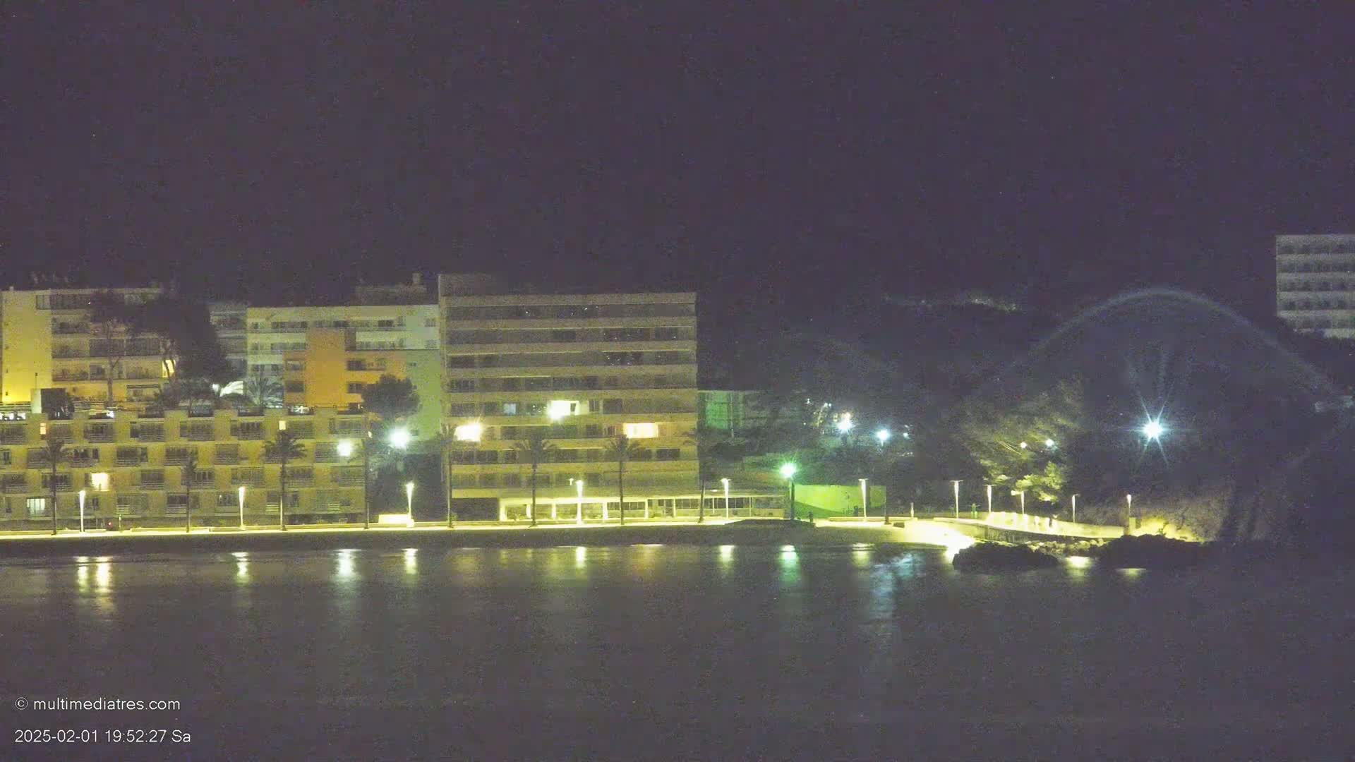 At night, illuminated apartment buildings line a calm, dark body of water under a clear, dark sky.