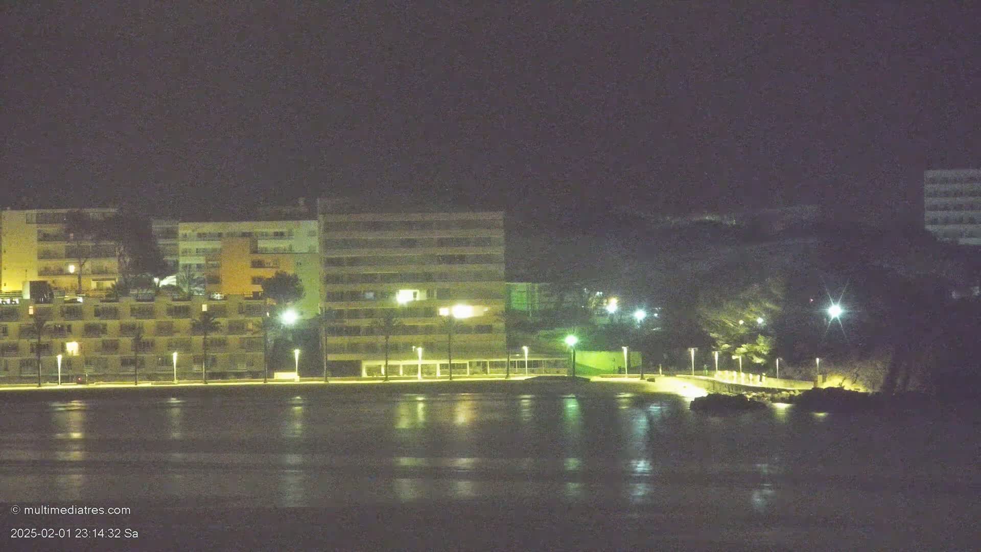 A nighttime waterfront scene shows several multi-story buildings illuminated by streetlights along a paved walkway, with dark water reflecting the lights in front.