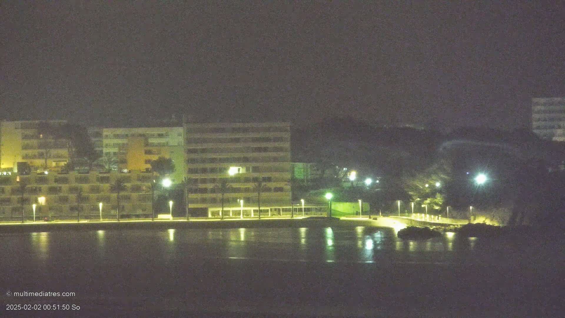 A nighttime view of multi-story buildings illuminated by streetlights along a waterfront, reflecting in calm water.