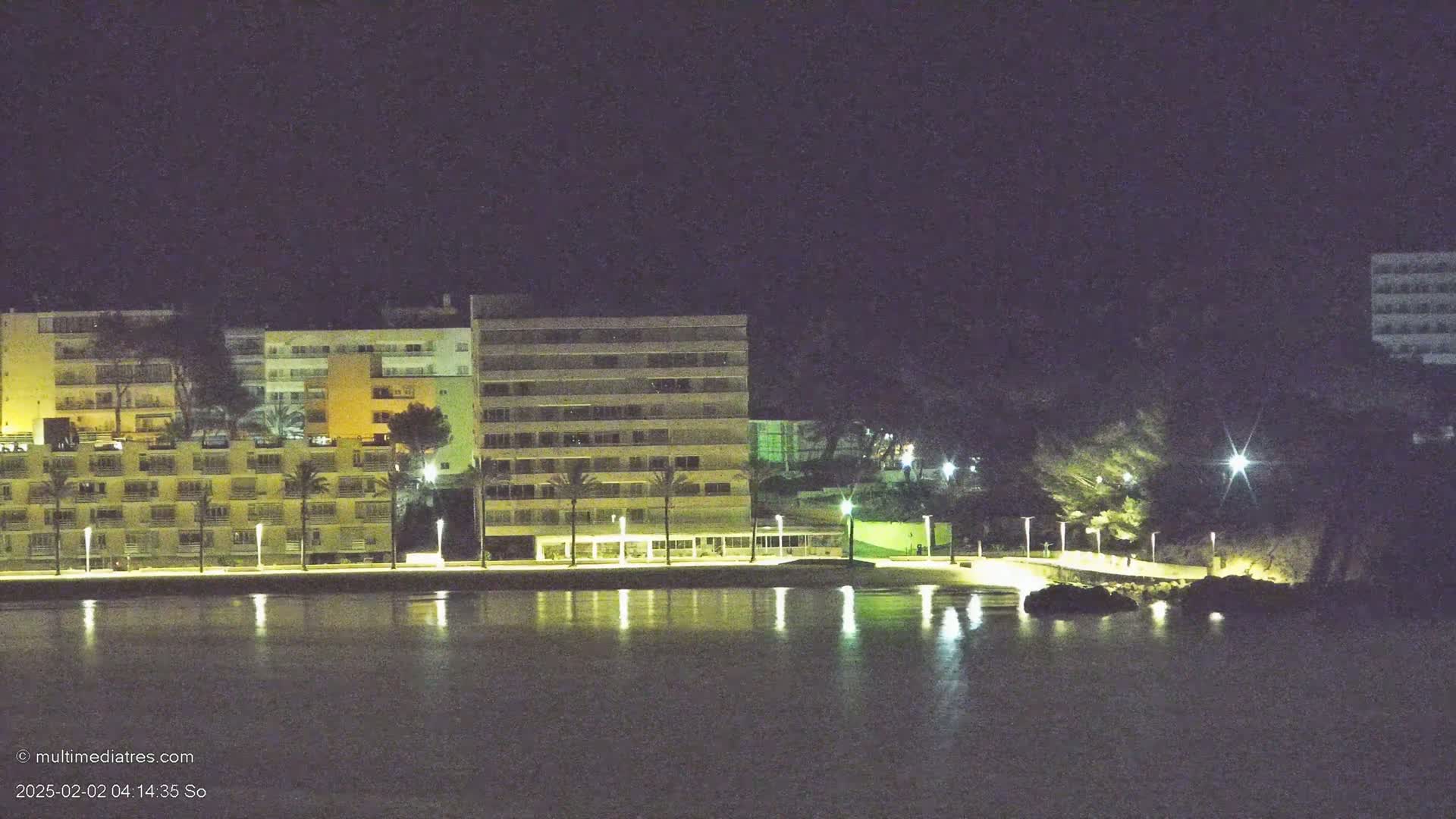 Illuminated multi-story buildings line a calm, dark body of water at night.