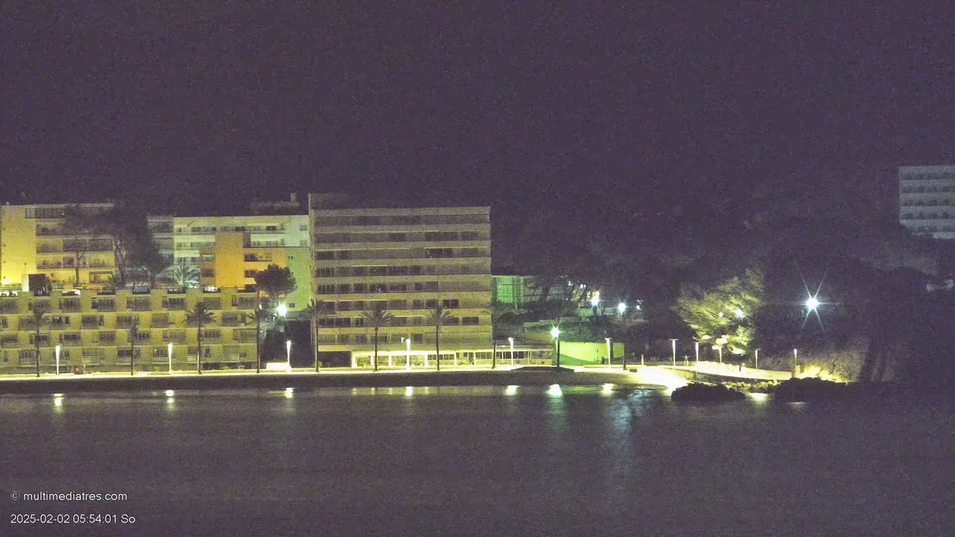 A waterfront area at night with several illuminated multi-story buildings, palm trees, and a calm body of water reflecting the lights.