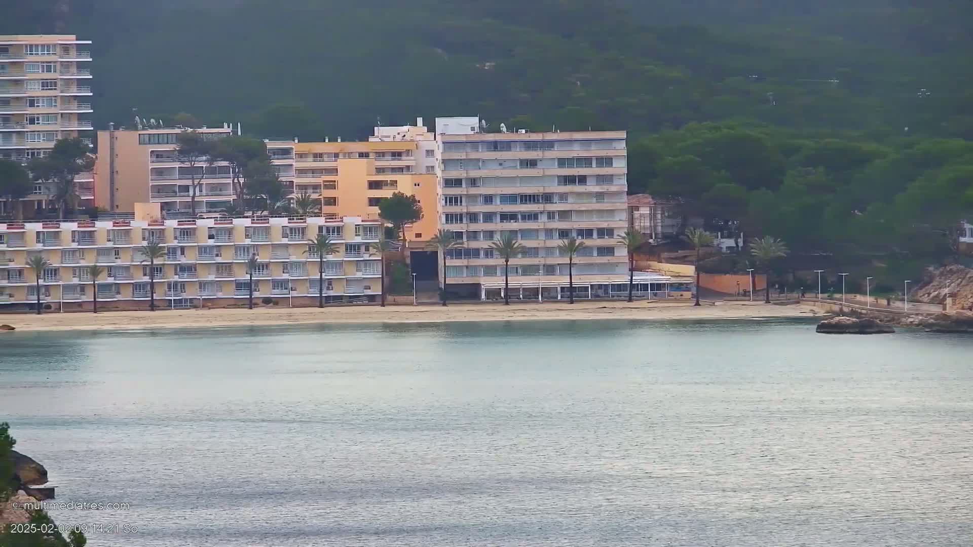 A calm body of water stretches out in front of several multi-story buildings situated on a sandy beach backed by a hill covered in dark green vegetation.