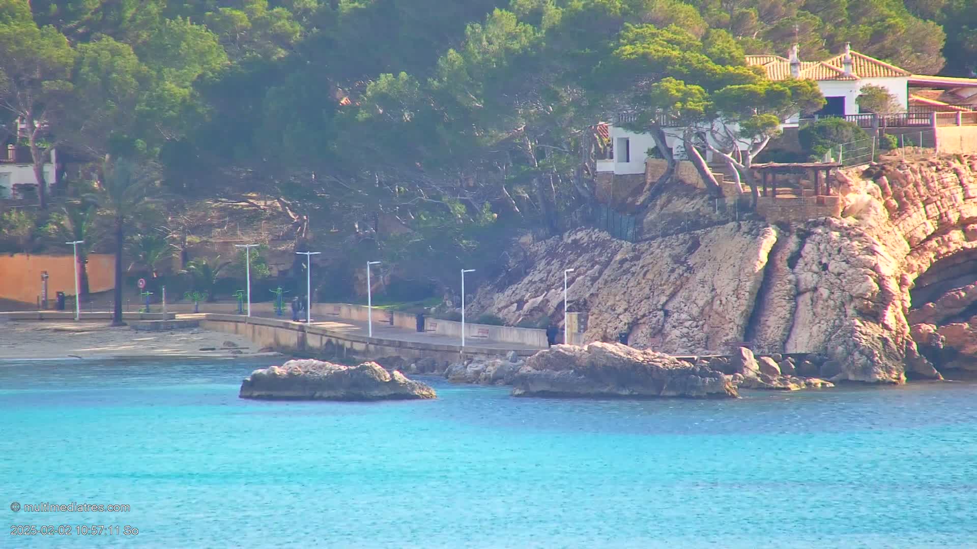 A rocky shoreline with a white building nestled among pine trees overlooks a calm, turquoise sea, with a paved walkway separating the beach from the water.