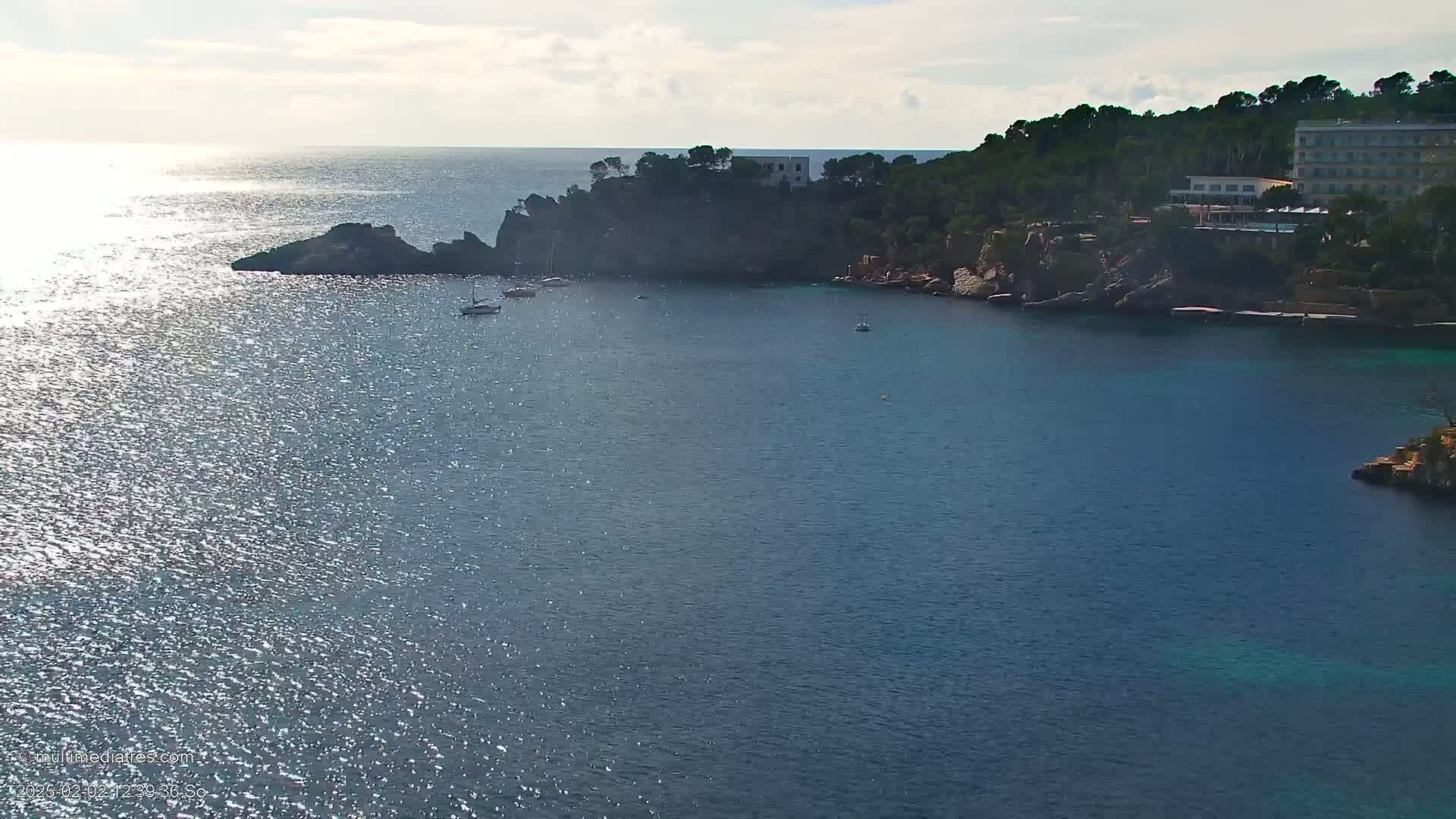 A tranquil cove with a few boats, a hotel, and rocky shores under a bright, sunny sky.