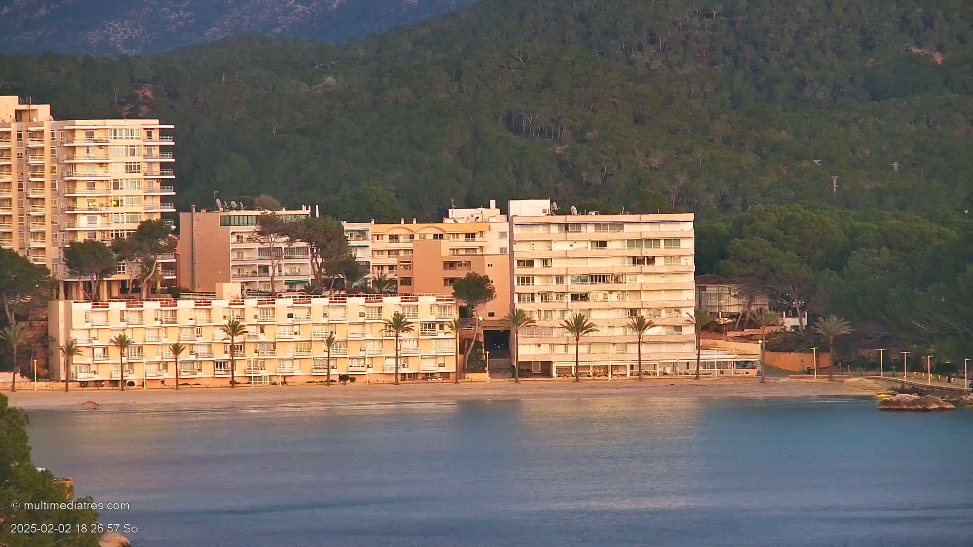 Several multi-story buildings line a sandy beach beside calm water, backed by a verdant hillside under a clear sky.
