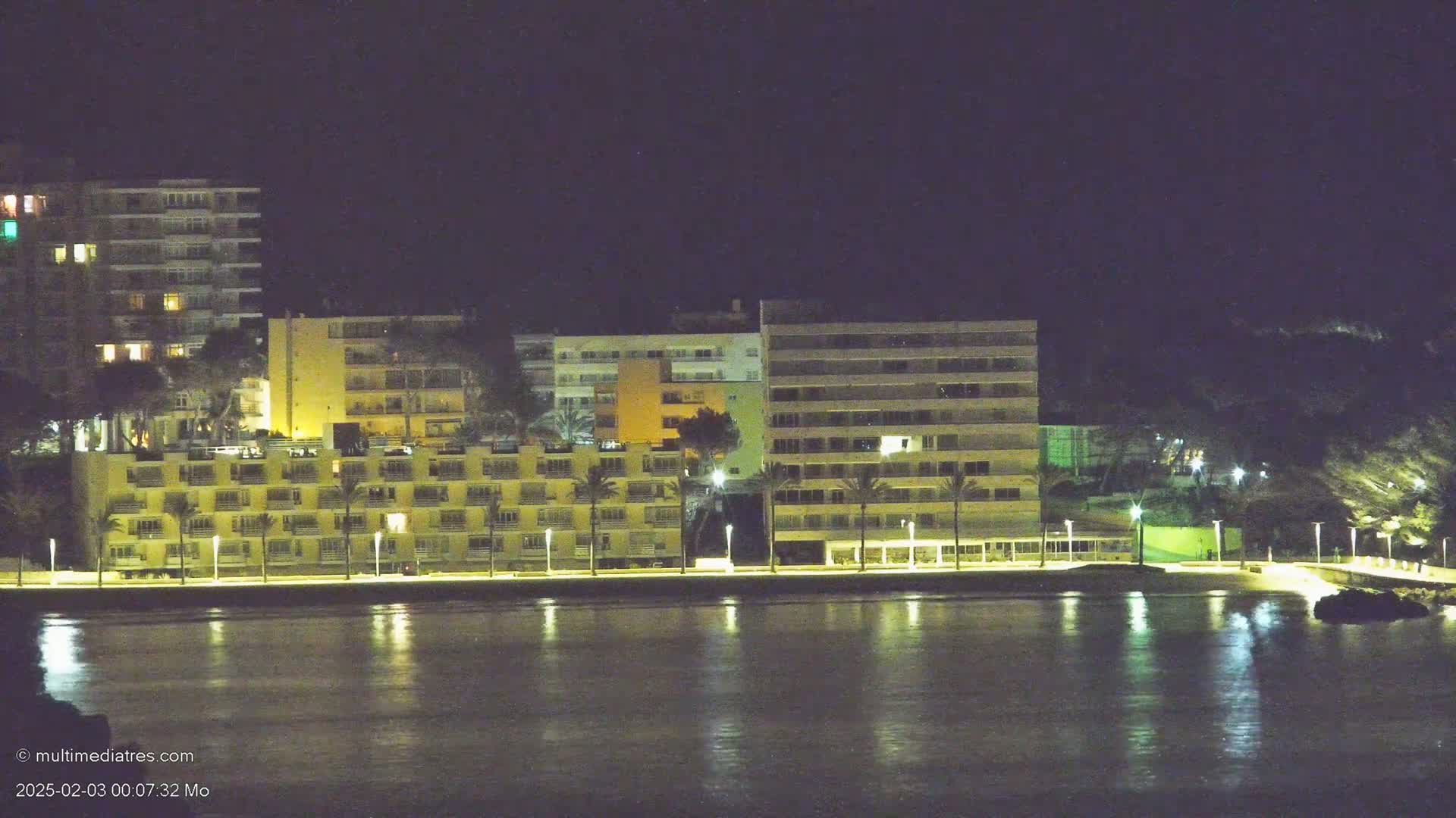 Illuminated apartment buildings line a waterfront at night, their lights reflecting on calm water.