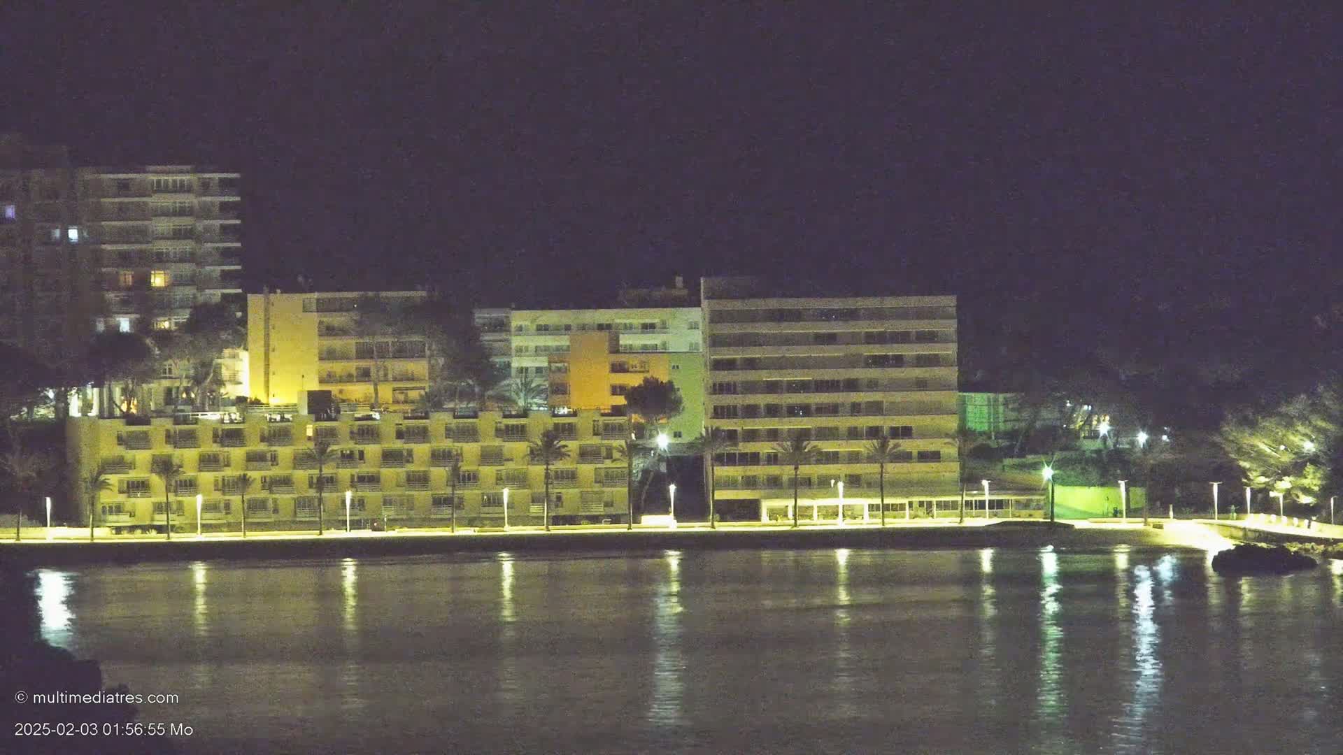 At night, several illuminated buildings line a waterfront, their lights reflecting on calm water.