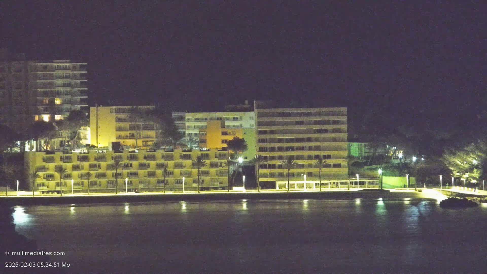 A row of illuminated beachfront buildings at night reflects in a calm body of water.