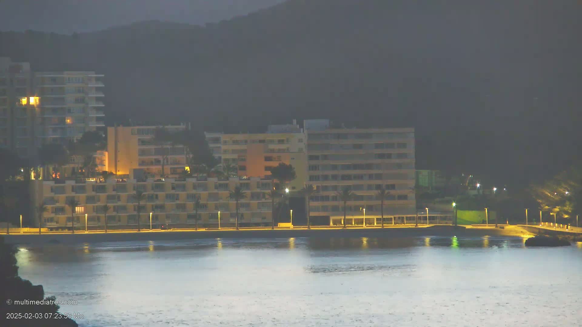 Lit-up apartment buildings line a calm, dark waterfront under an overcast night sky.