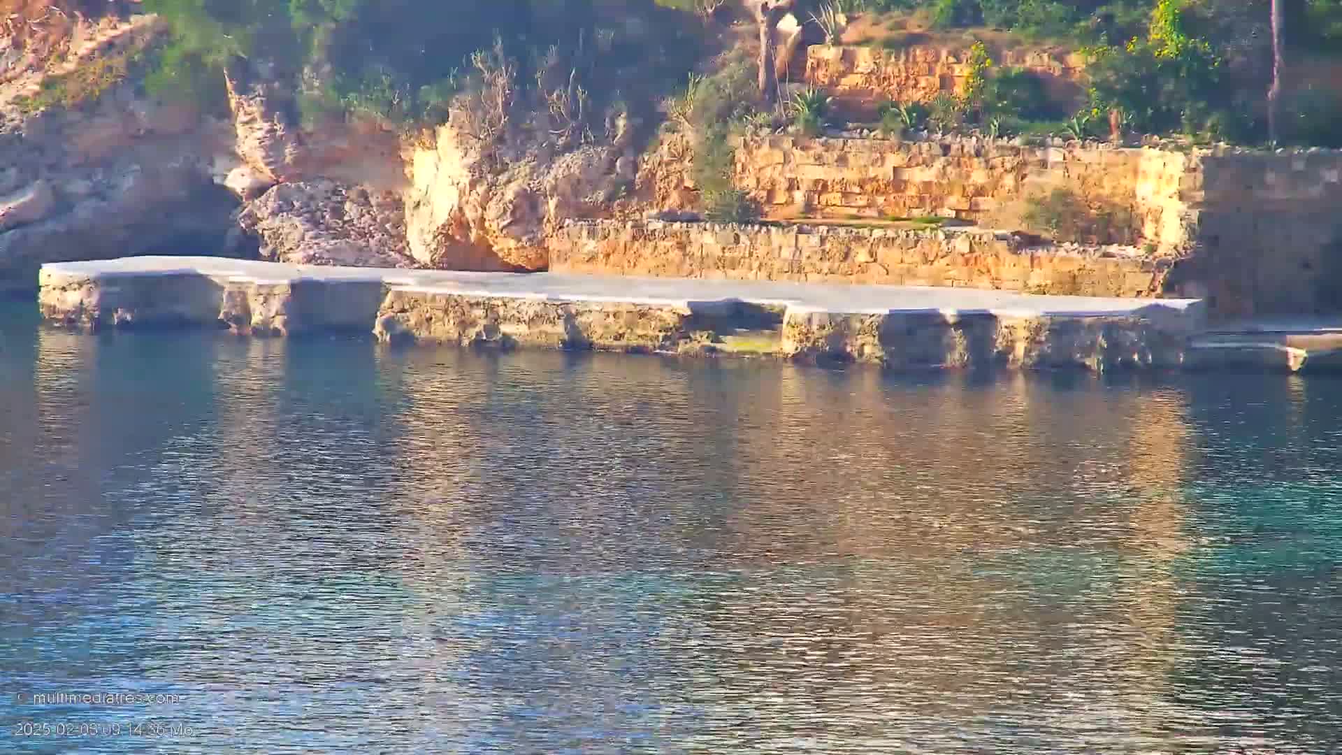 A calm body of water reflects a rocky shoreline with a flat, paved area and stone retaining walls.