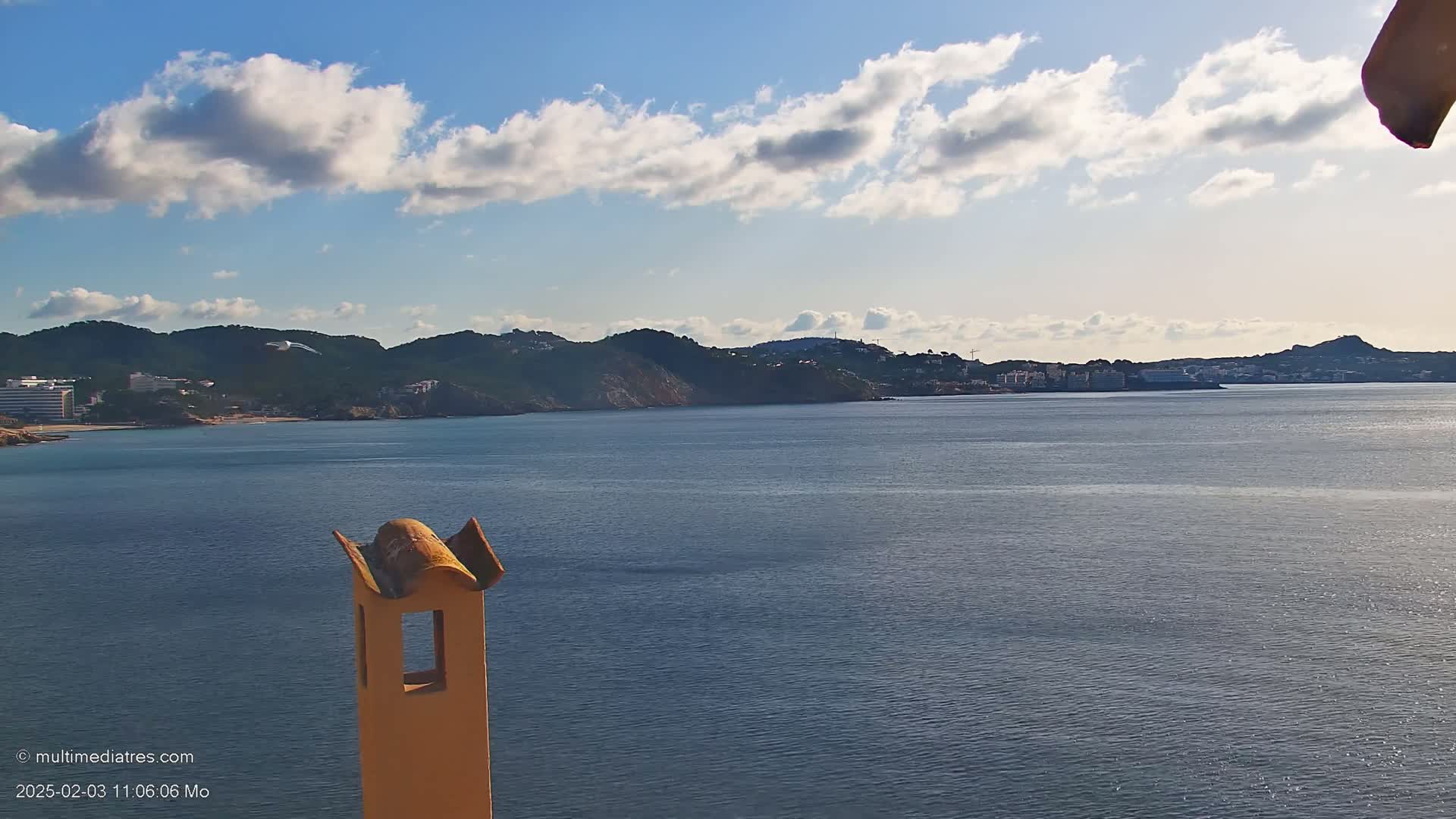 A partly cloudy sky overlooks a calm sea separating a beach and coastal buildings from distant hills.