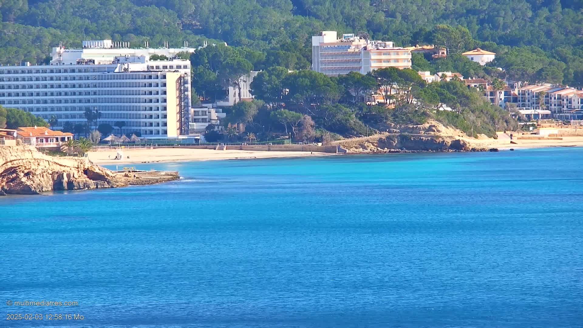 A sandy beach with turquoise water is nestled between a hillside of green trees and several large hotel buildings.