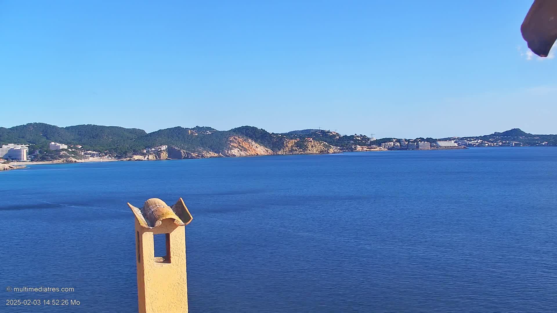A tan chimney stands in the foreground against a calm, blue ocean with a distant coastline of green hills and buildings under a clear blue sky.