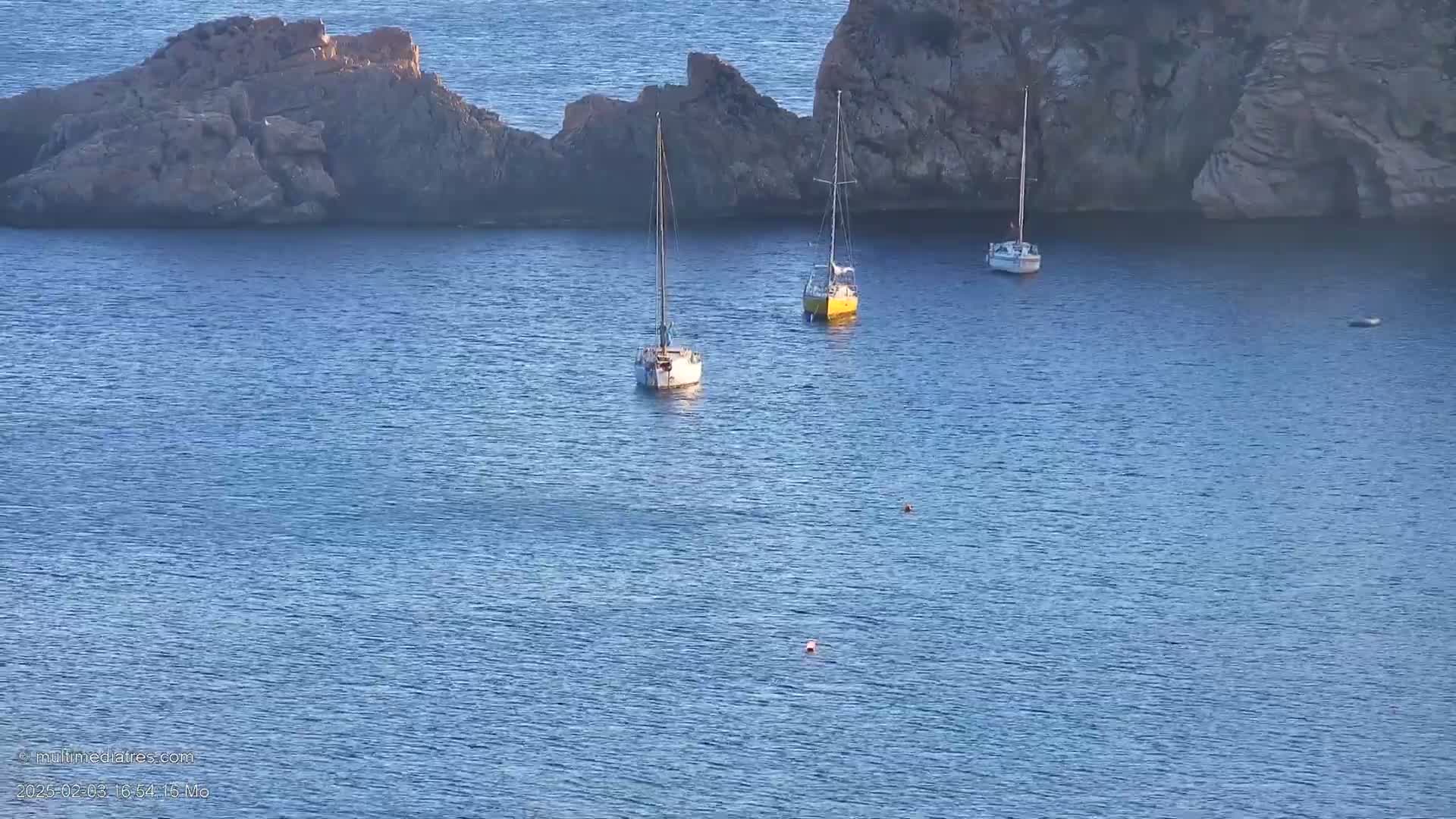 Three sailboats are anchored in calm blue water near a rocky shoreline.