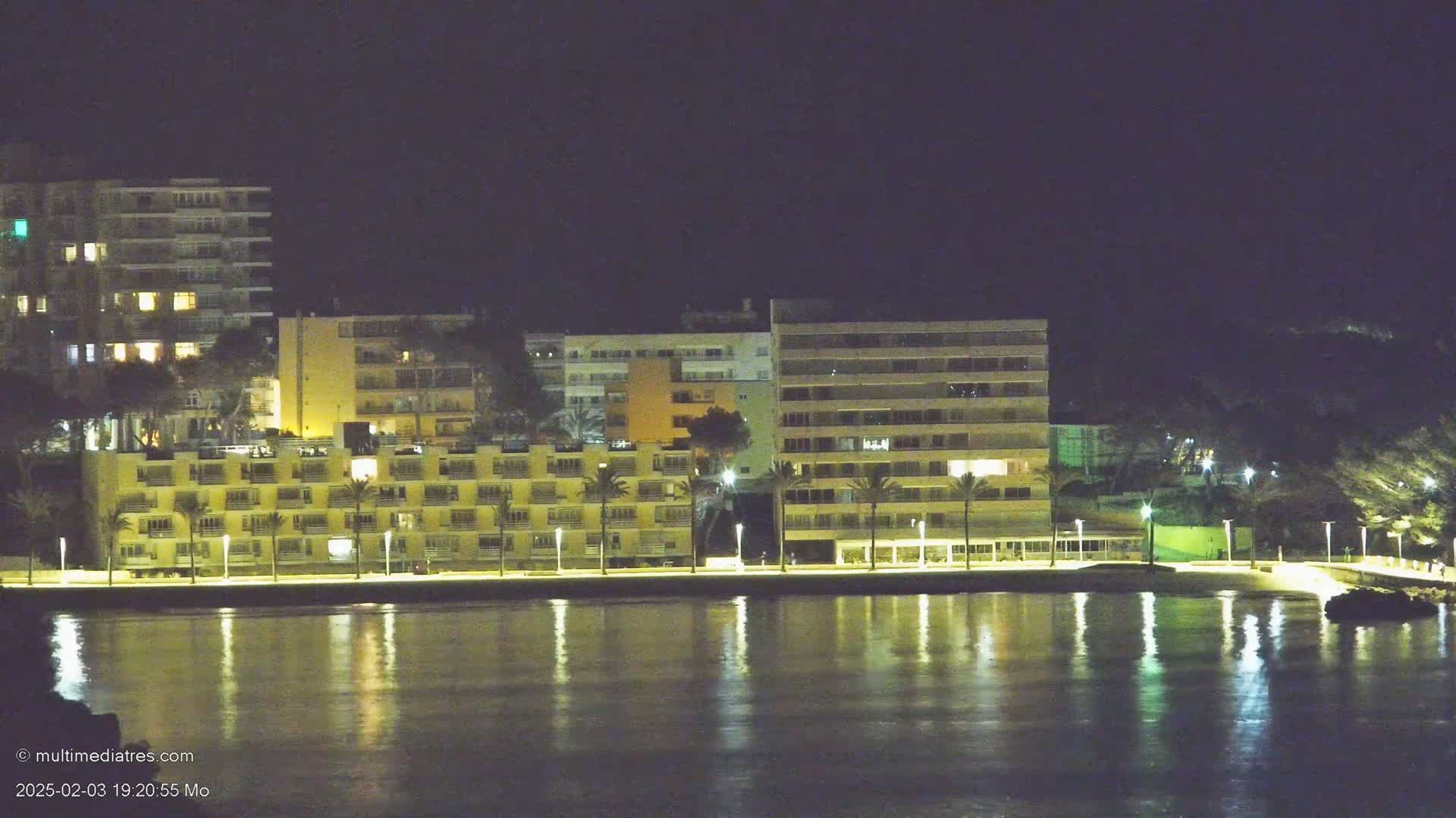 At night, illuminated buildings line a waterfront, their reflections shimmering on calm water.