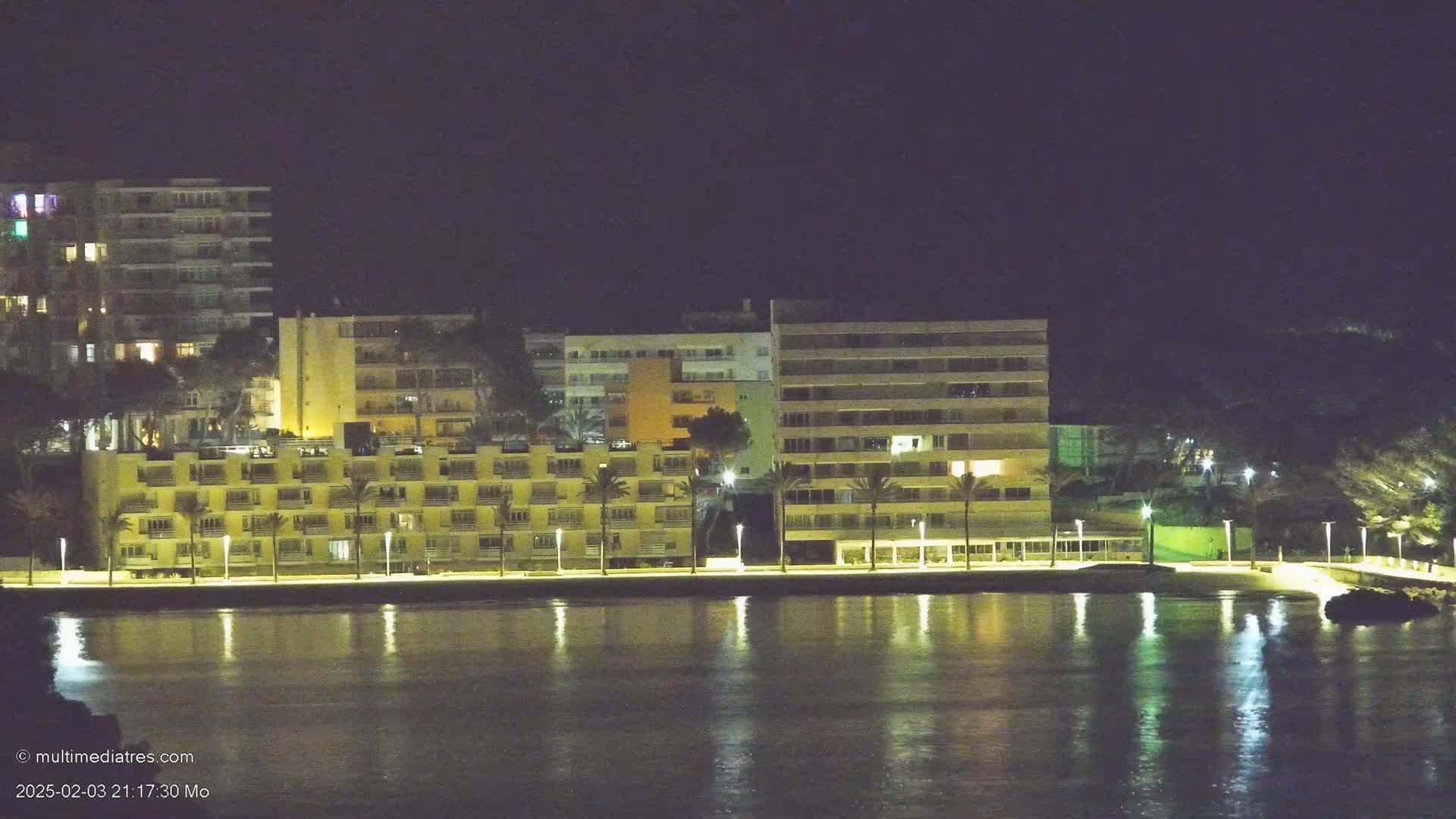 A waterfront of multi-story buildings is lit up at night, reflected in calm water.