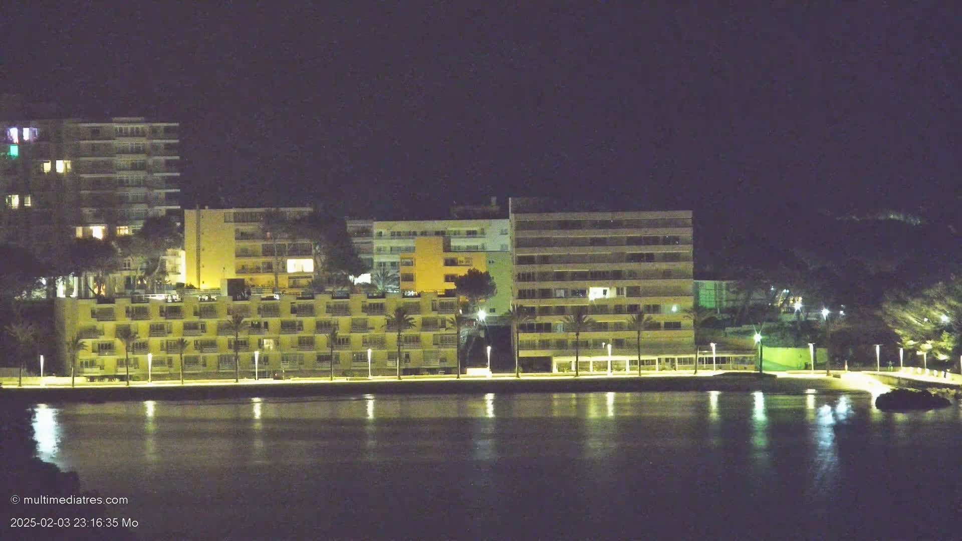 A row of illuminated apartment buildings lines a calm body of water at night.