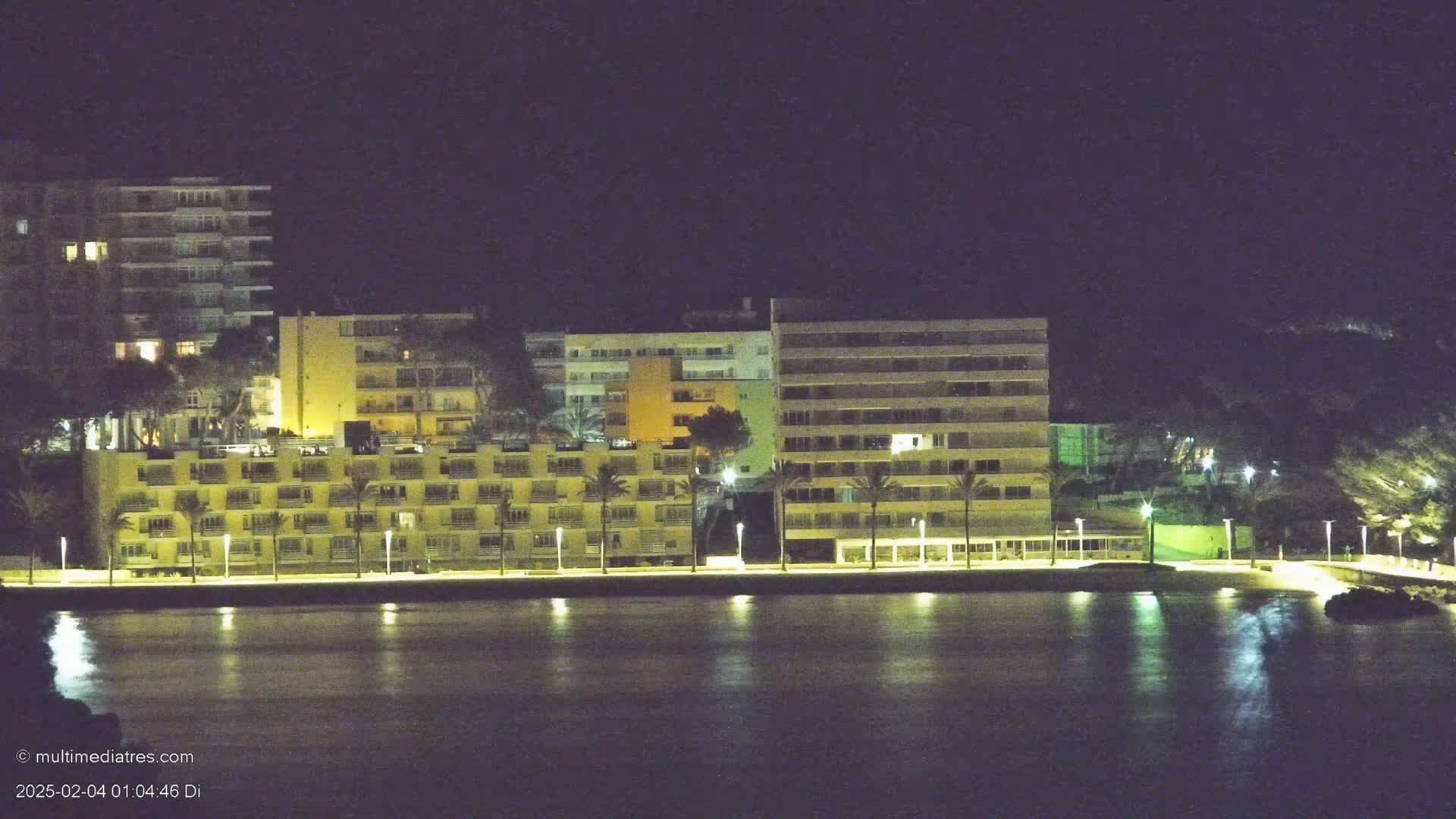 A row of illuminated beachfront buildings at night, reflected in calm water.