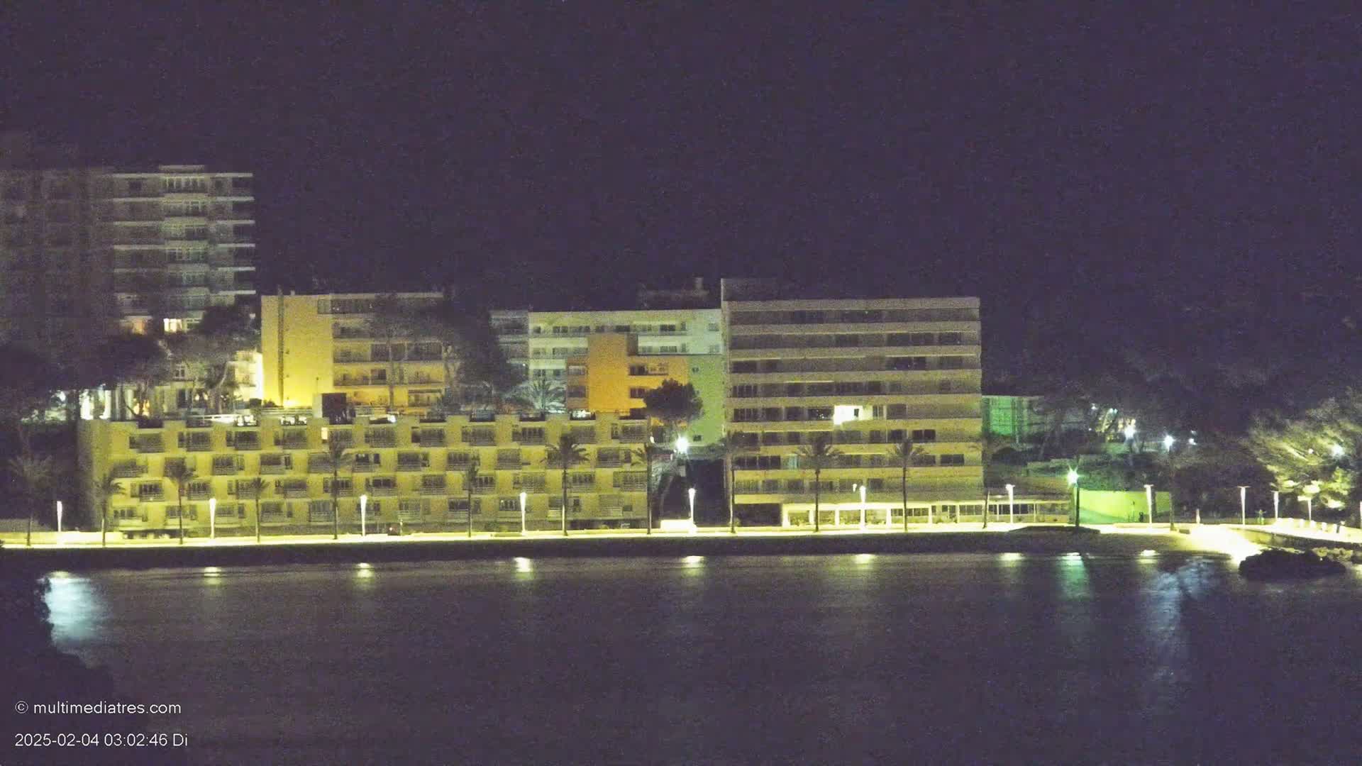 A row of illuminated multi-story buildings stands along a waterfront at night.