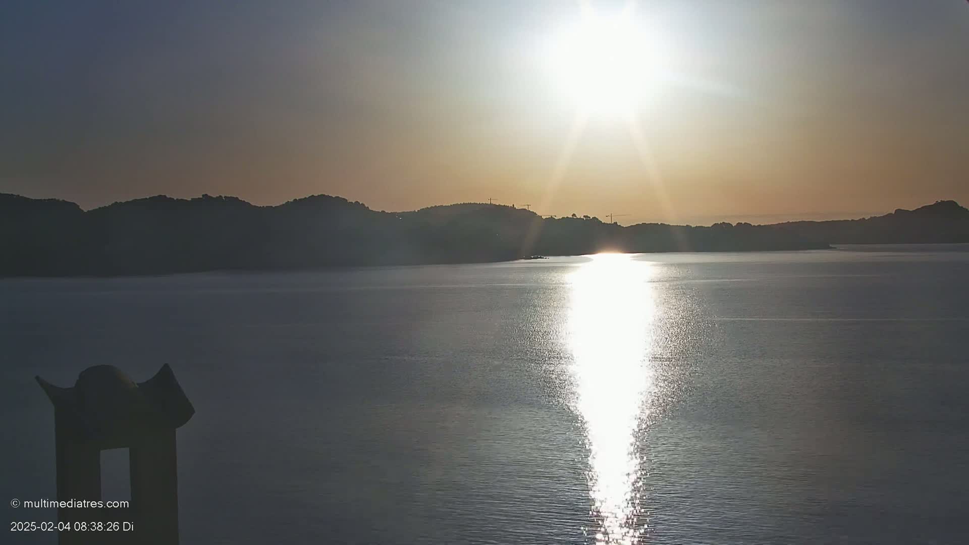 A calm body of water reflects the bright sun shining over a dark, hilly coastline at sunrise.