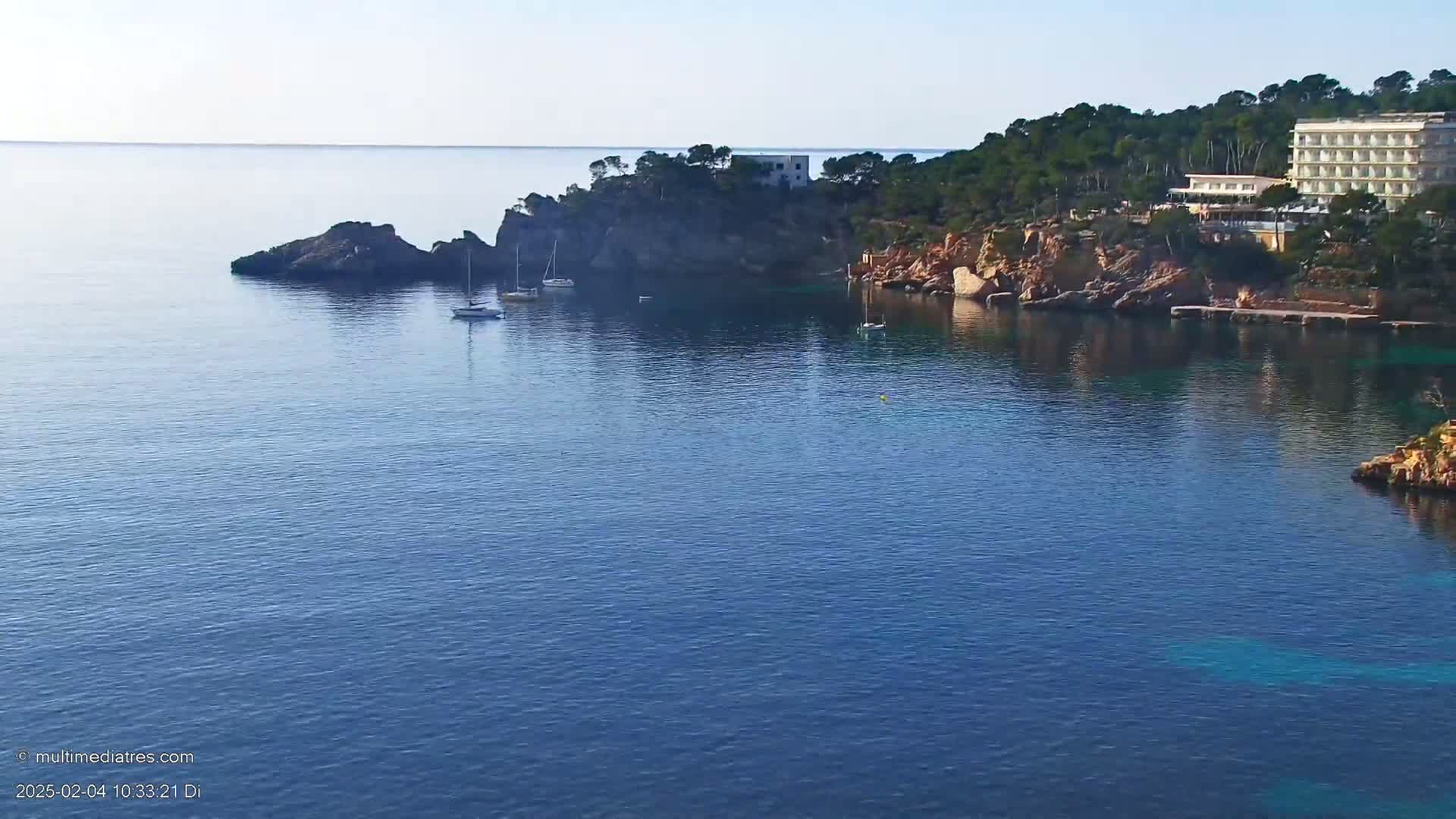 A calm, blue ocean cove is nestled between rocky cliffs, with a few sailboats anchored nearby and a hotel situated on the hillside.