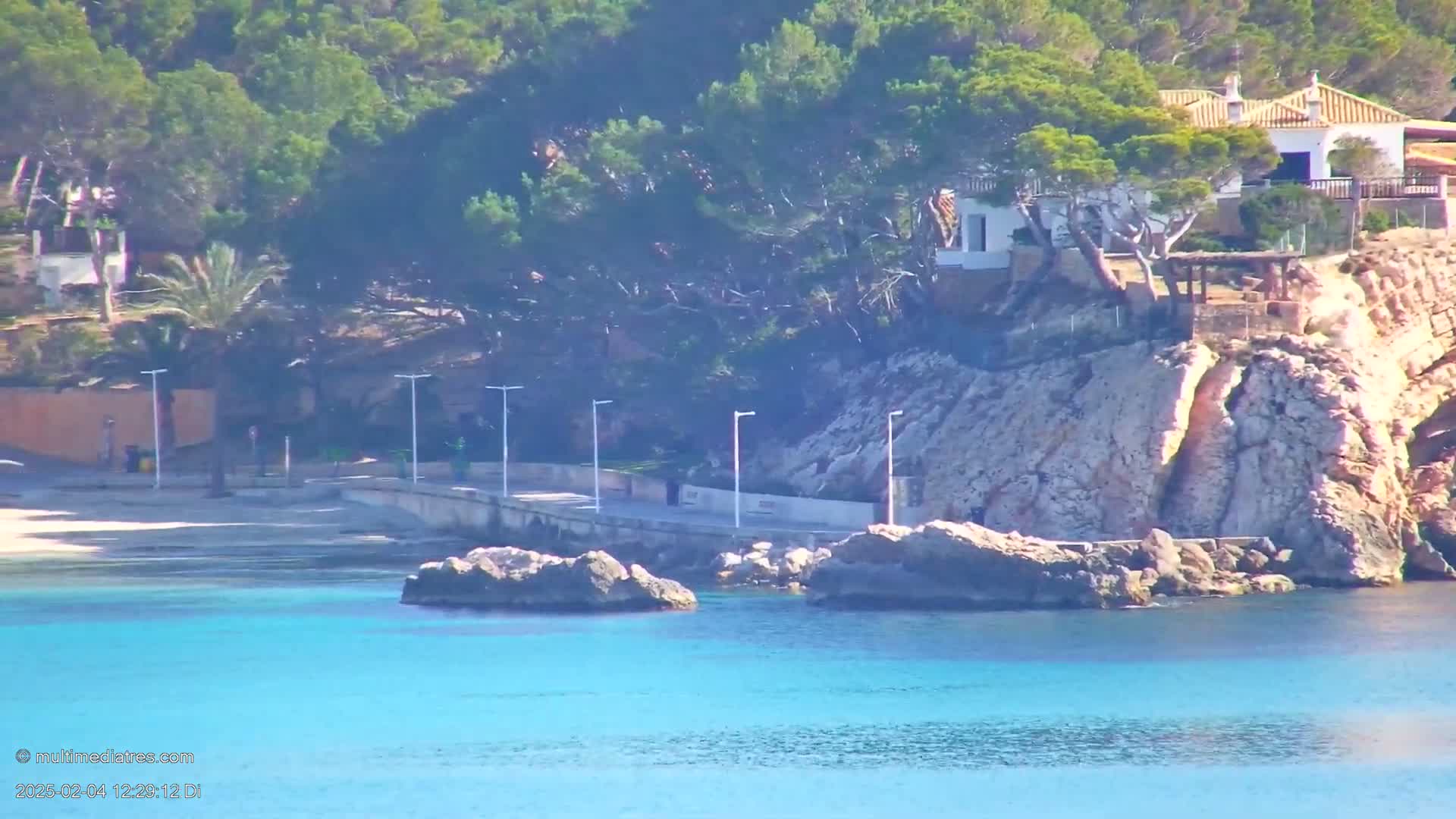 A white house sits atop a rocky cliff overlooking a calm, bright blue body of water with a small sandy beach and a paved walkway.