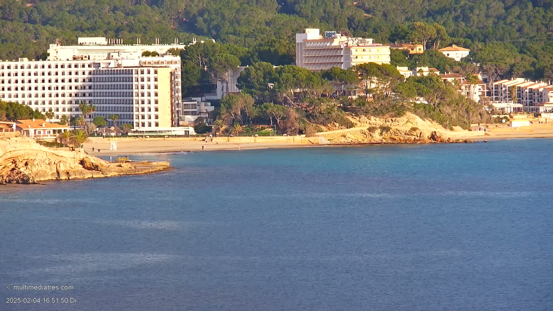 A sandy beach with calm, clear turquoise water lies in front of several hotels and buildings nestled amongst a hillside covered in green vegetation.