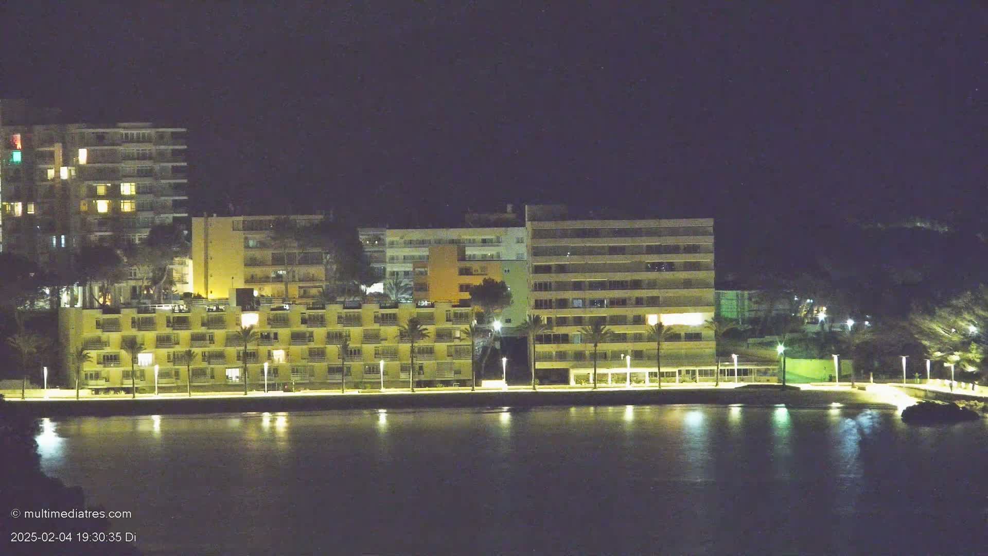 A row of illuminated beachfront buildings at night, reflected in calm water.