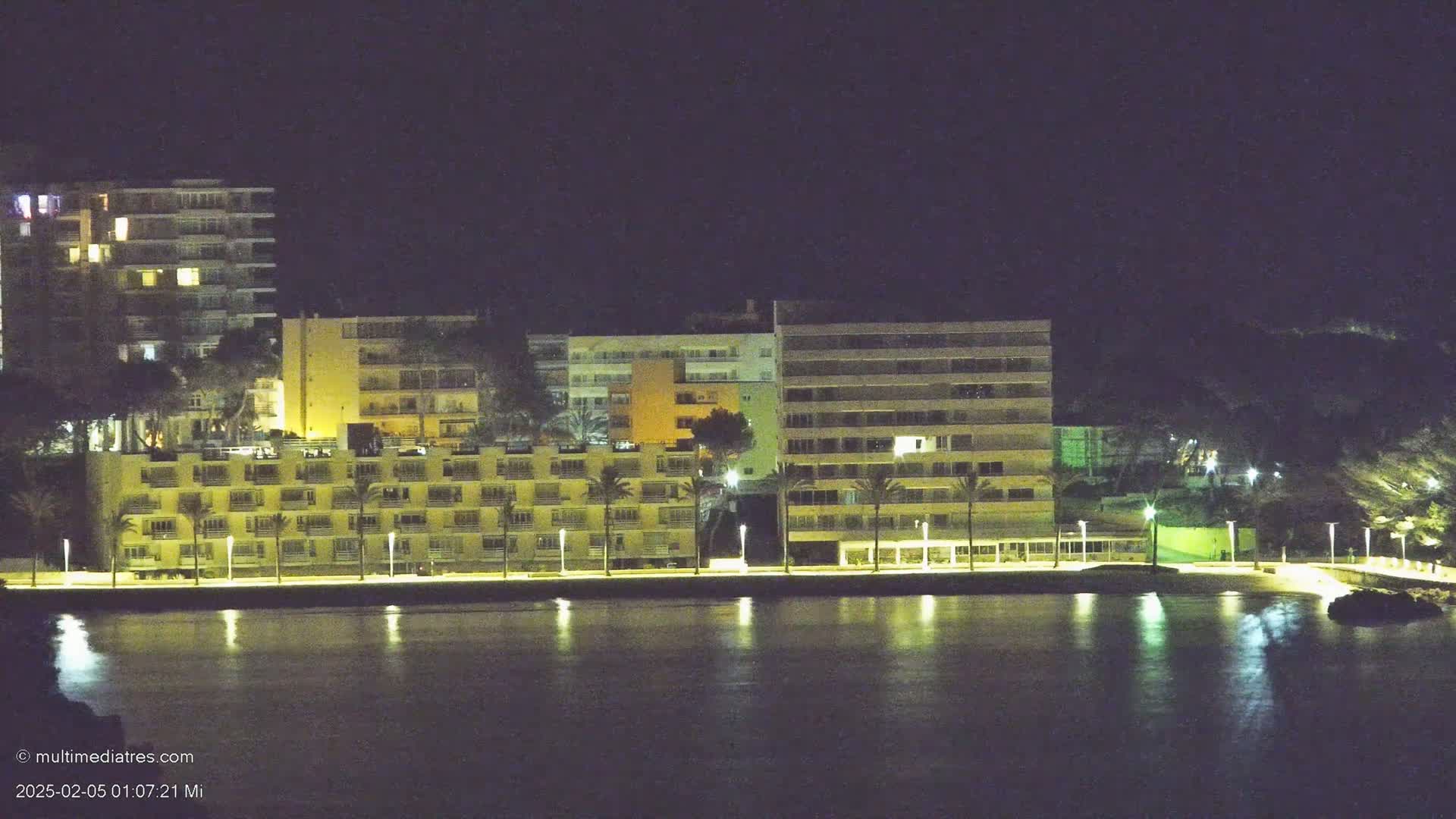 Illuminated buildings line a waterfront at night, reflected in calm water.