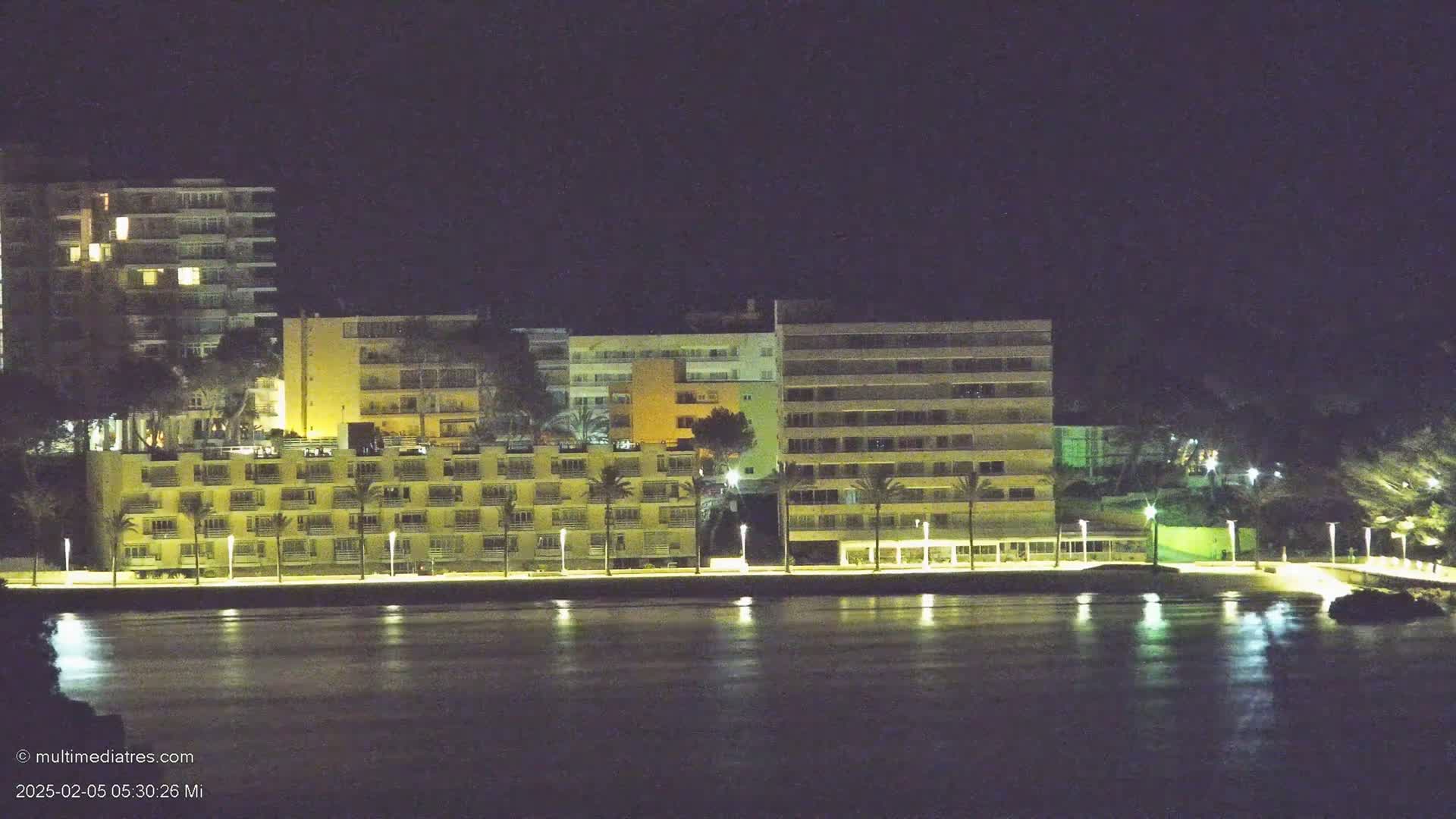 A row of illuminated buildings along a waterfront at night, reflected in calm water.