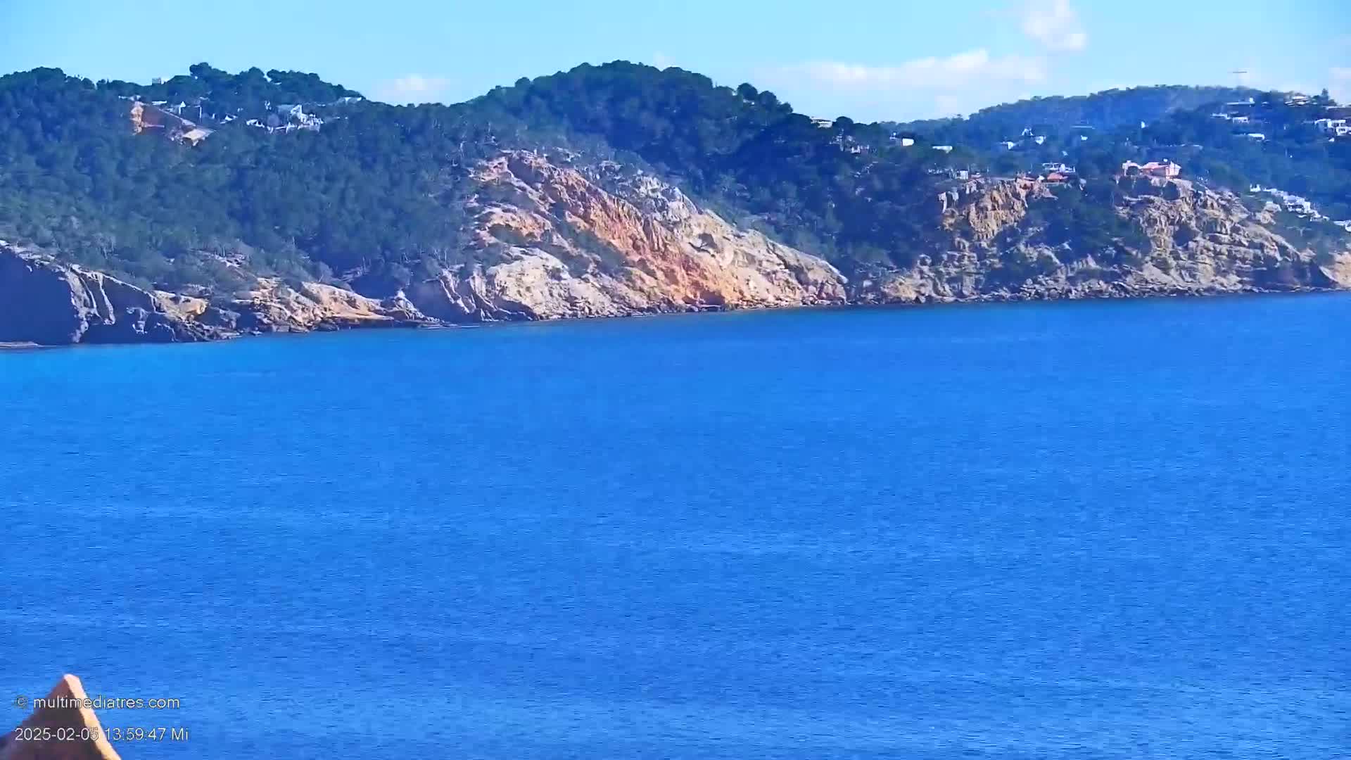 A rocky, tree-covered hillside with houses on top overlooks a calm, deep blue ocean under a clear sky.