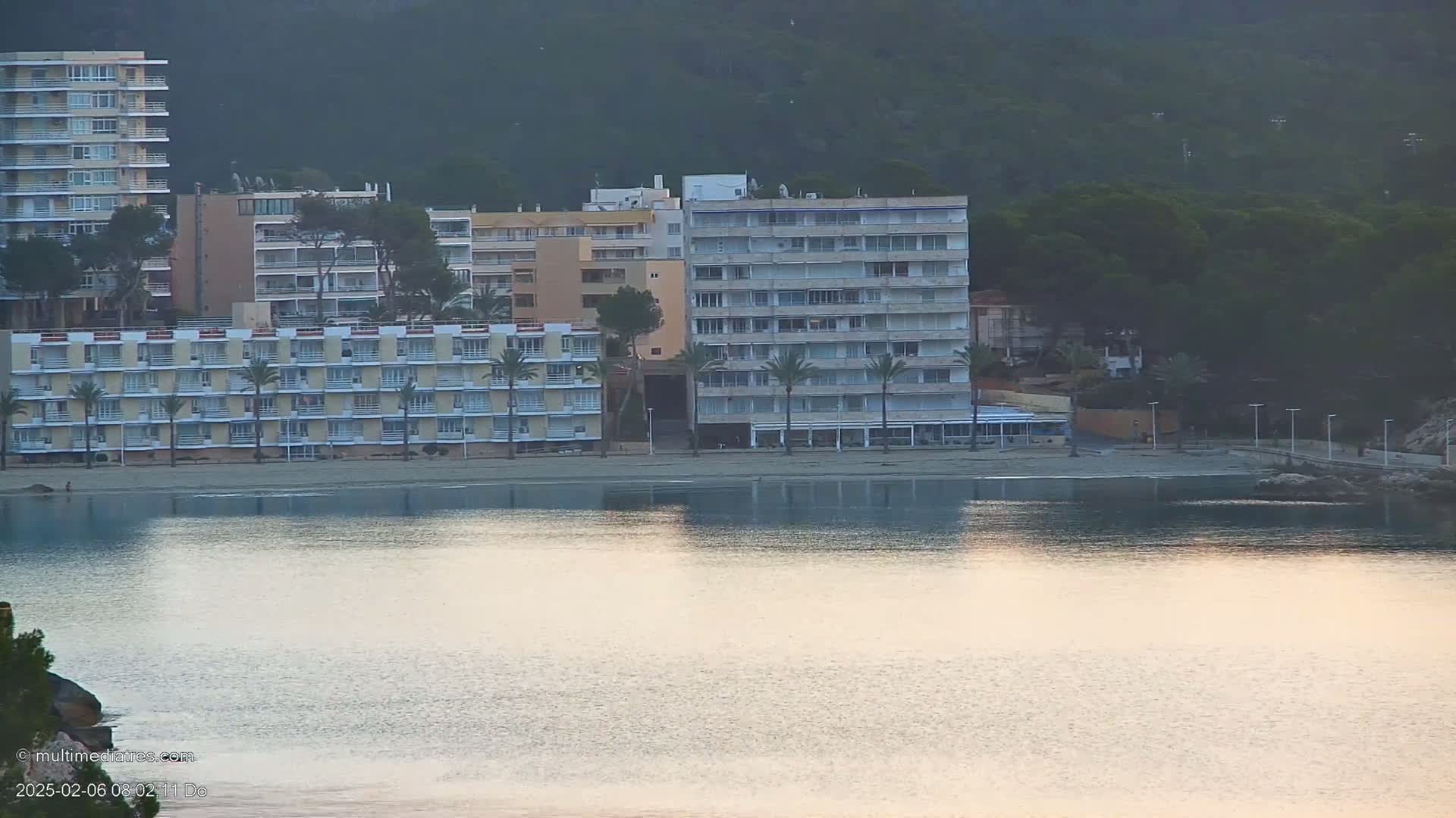 Several multi-story buildings line a sandy beach next to calm, reflective water under a dusky sky.