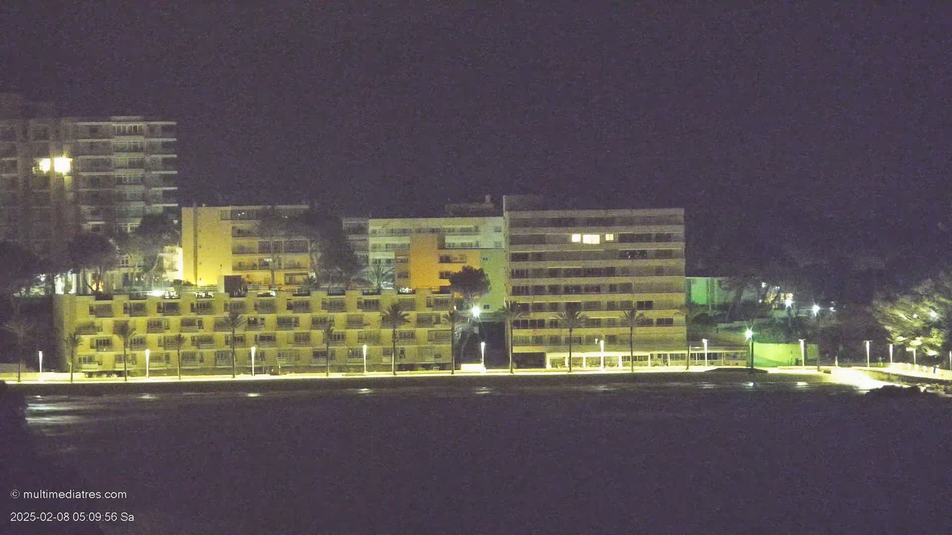 A row of illuminated buildings, some multi-story hotels, sits along a dark waterfront at night.