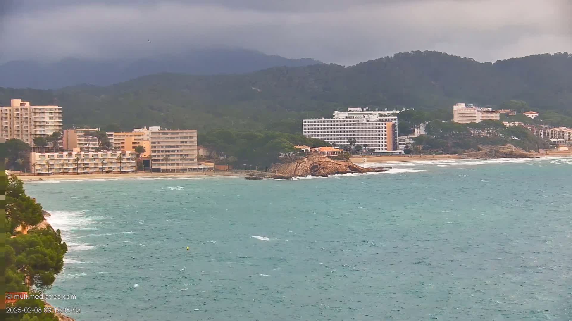 A coastal scene shows several high-rise hotels and buildings along a sandy beach with a rocky outcropping in the water, under an overcast sky with a choppy sea.