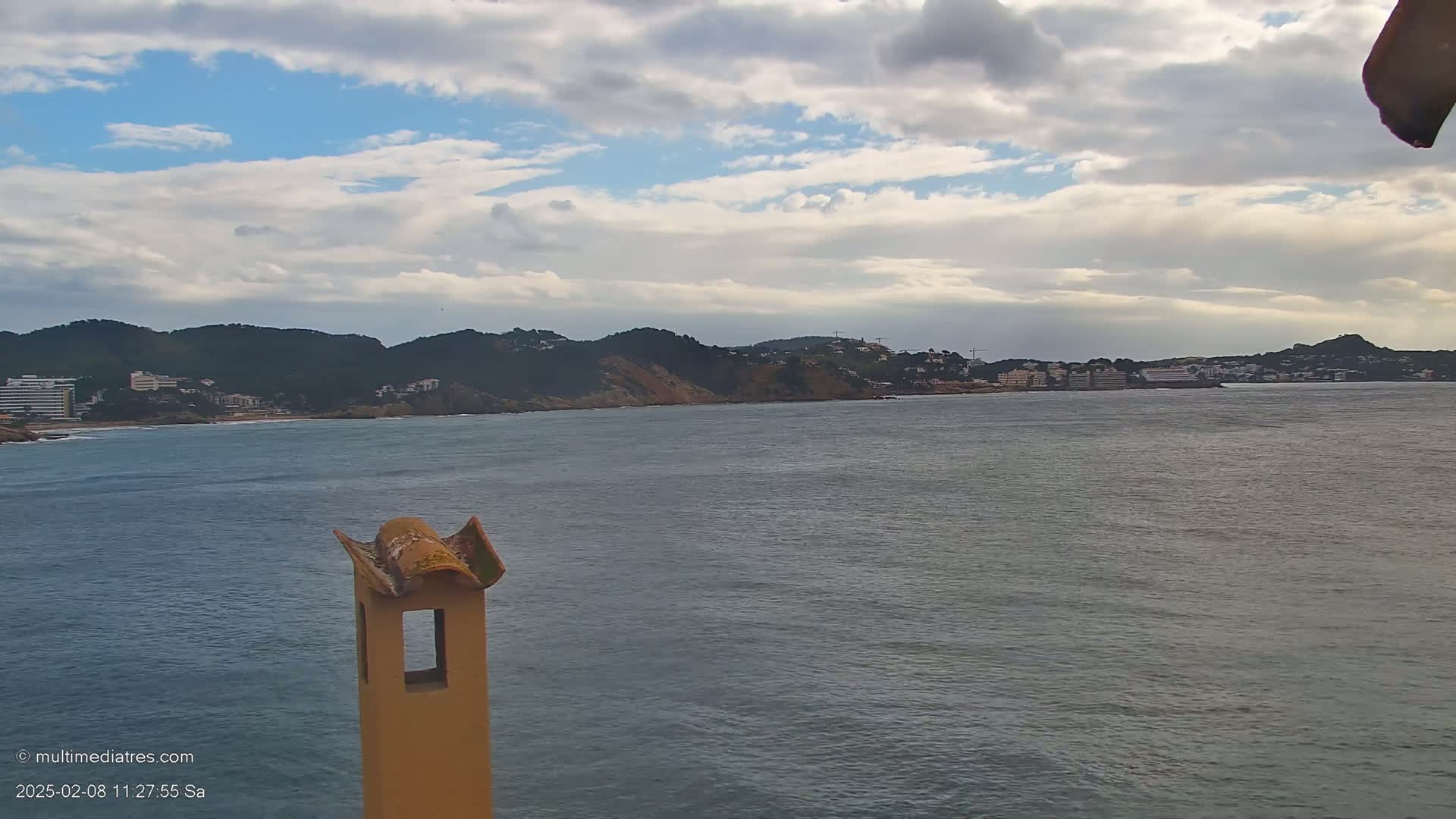 A partly cloudy day reveals a coastal scene of a calm ocean with buildings and hills in the distance, viewed from behind a tan chimney.