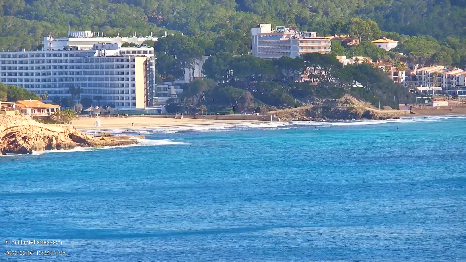 A sandy beach with turquoise water laps against a rocky shore, backed by hotels and buildings nestled among green hills under a sunny sky.
